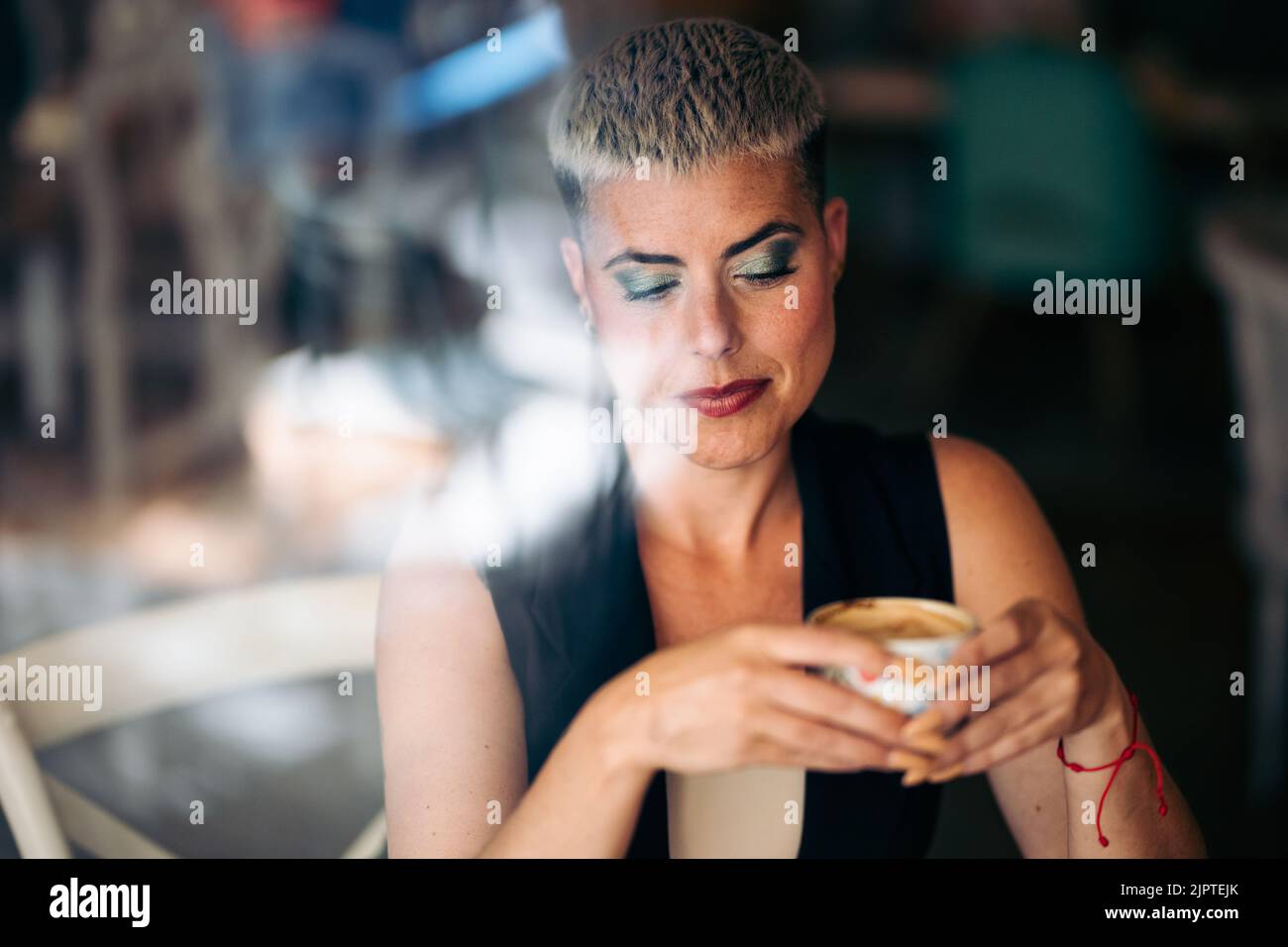 Nachdenkliche Frau mit kurzen Haaren, die einen Kaffee von außerhalb eines Fensters hält Stockfoto