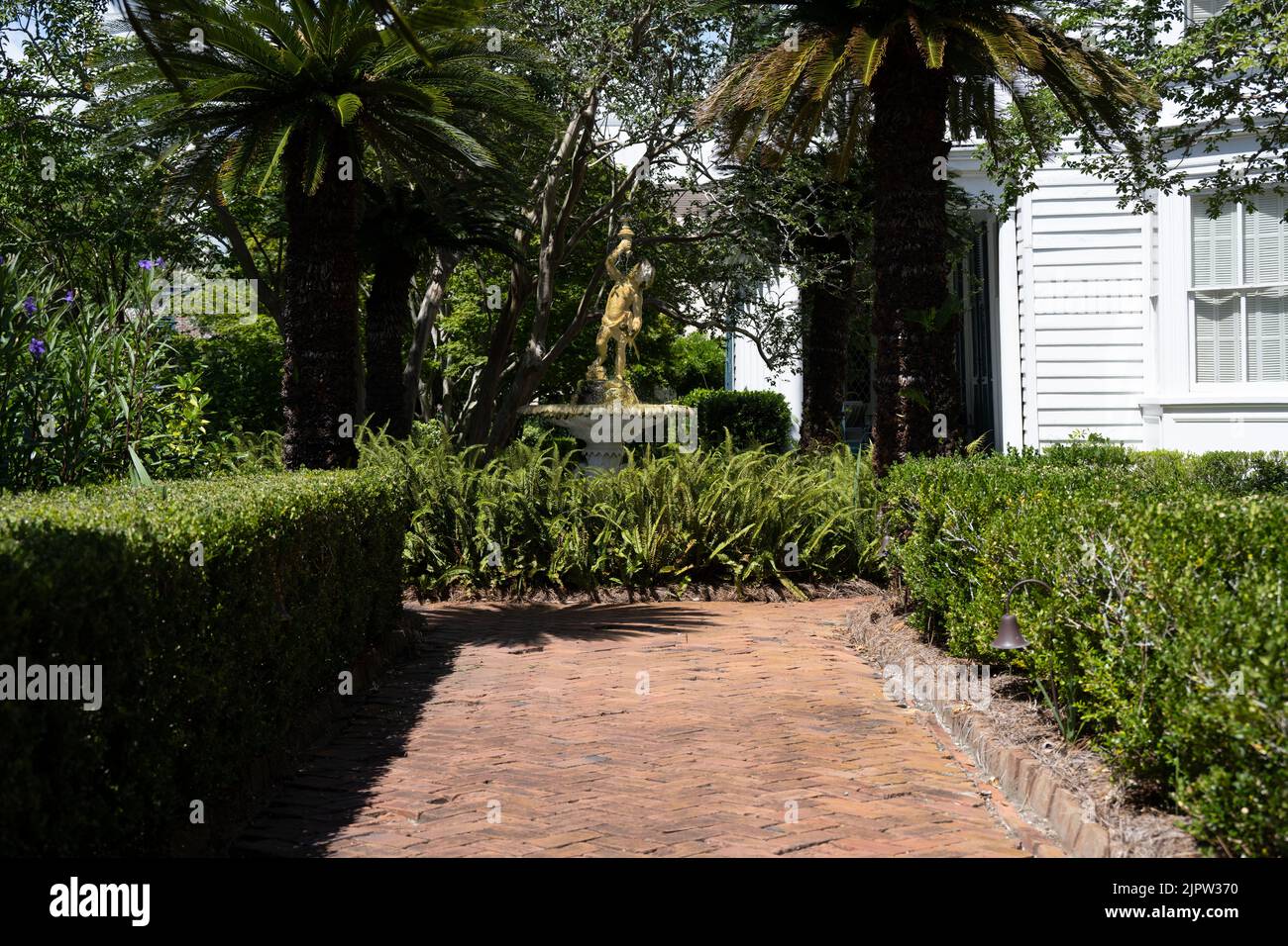 Eine Skulptur aus einem weißen Brunnen hinter einem roten Backsteinweg, der von Hecken umgeben ist Stockfoto