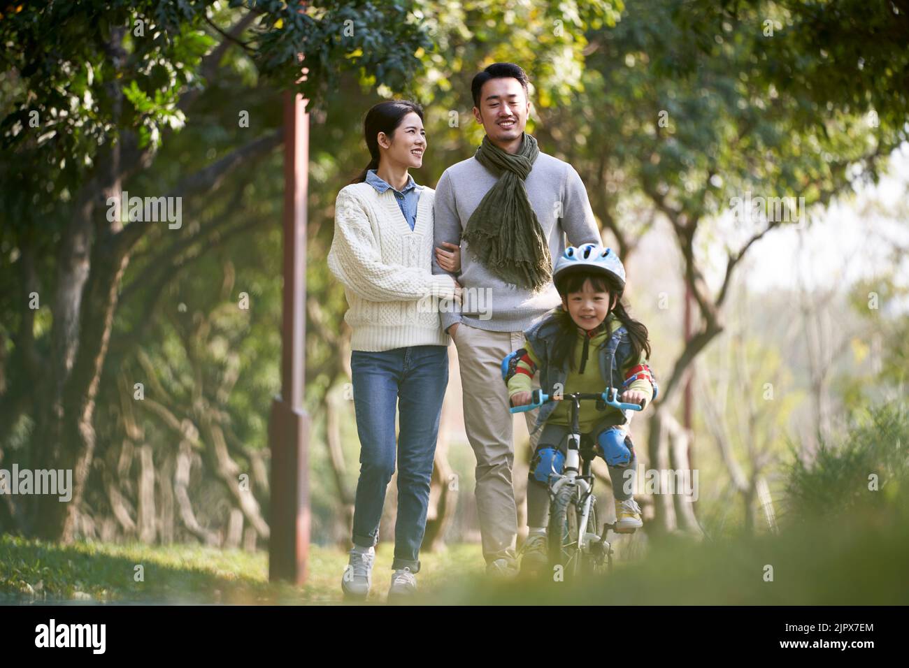 Junges asiatisches Paar, das im Park spazieren geht, während Tochter glücklich und lächelnd auf einem Kinderfahrrad reitet Stockfoto