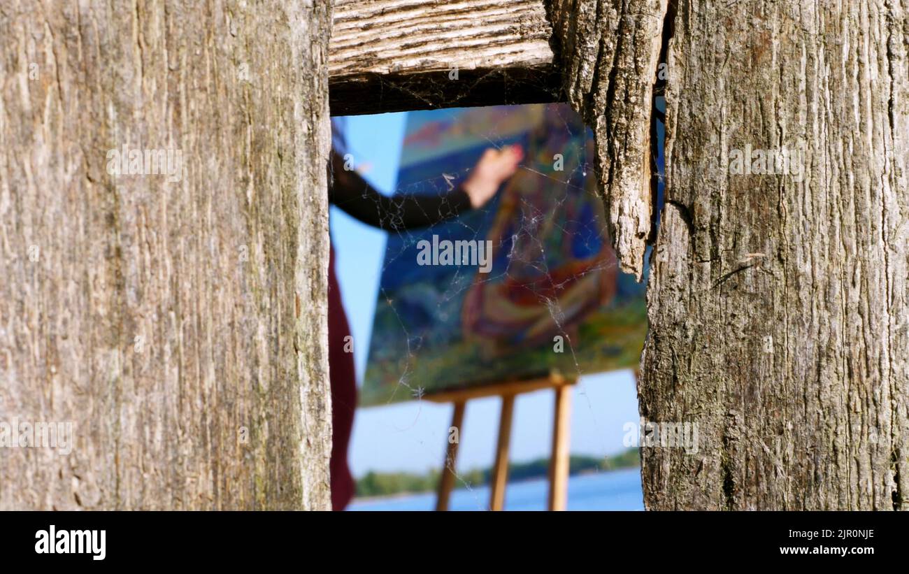 Durch einen Riss in einem Holzzaun, bedeckt mit Spinnweben, kann man die Hand mit dem Pinsel der Künstlerin sehen, sie malt auf Leinwand den gott Shiva. Hochwertige Fotos Stockfoto