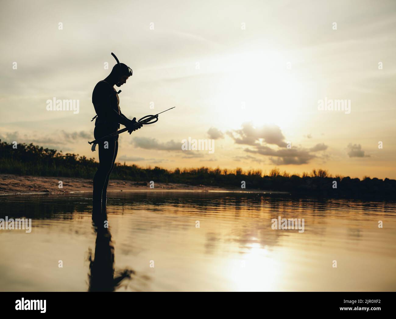 Seitenansicht des Speerfischers, der seine Speerpistole kontrolliert, während er im Meerwasser steht. Abenteuerlicher junger Mann, der bei Sonnenuntergang Speerfischen geht. Stockfoto