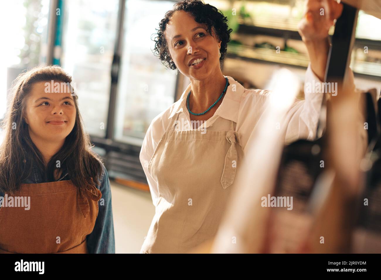 Fröhliche Geschäftsleiterin, die ihrem neuen Mitarbeiter in einem Lebensmittelgeschäft Anweisungen gibt. Weibliche Kleinunternehmerin, die eine Frau mit Down-Syndrom beschäftigt Stockfoto