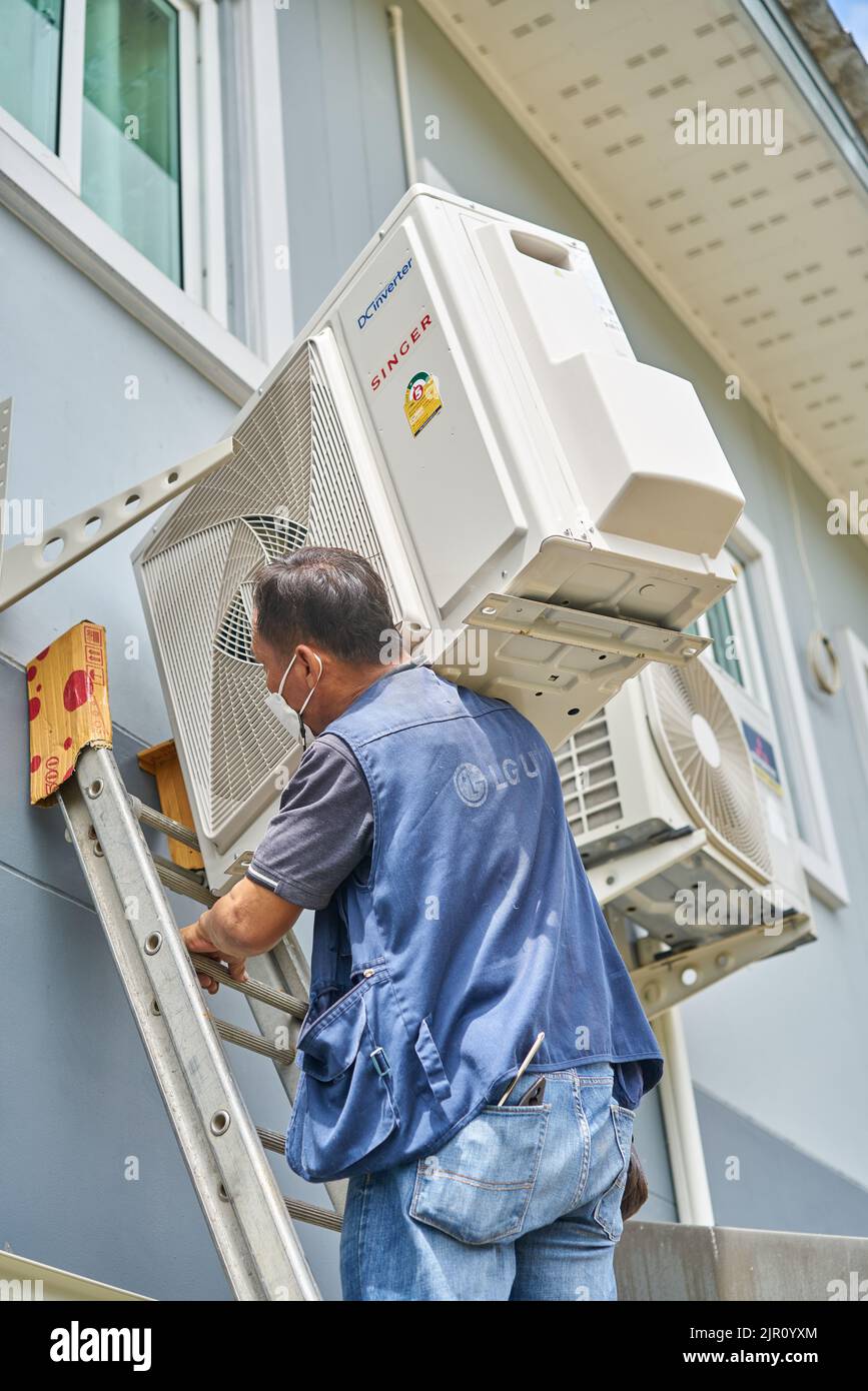 Ein Mann trägt einen schweren Klimaanlagenkompressor auf seiner Schulter, während er eine Leiter klettert. Stockfoto