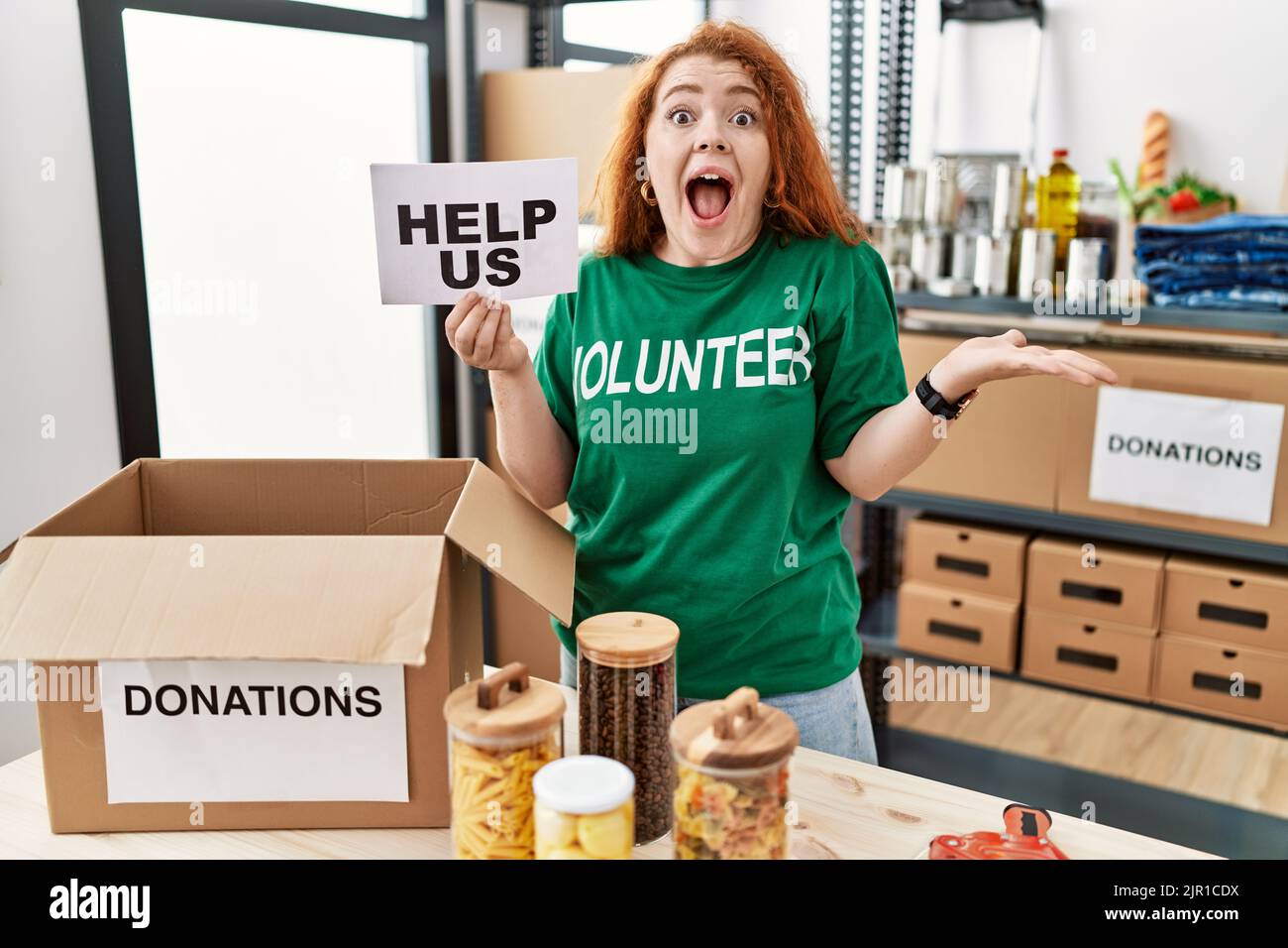 Junge Rotschopf Frau trägt Freiwilligen T-Shirt halten helfen uns Banner feiern Sieg mit glücklichem Lächeln und Sieger Ausdruck mit erhobenen Händen Stockfoto