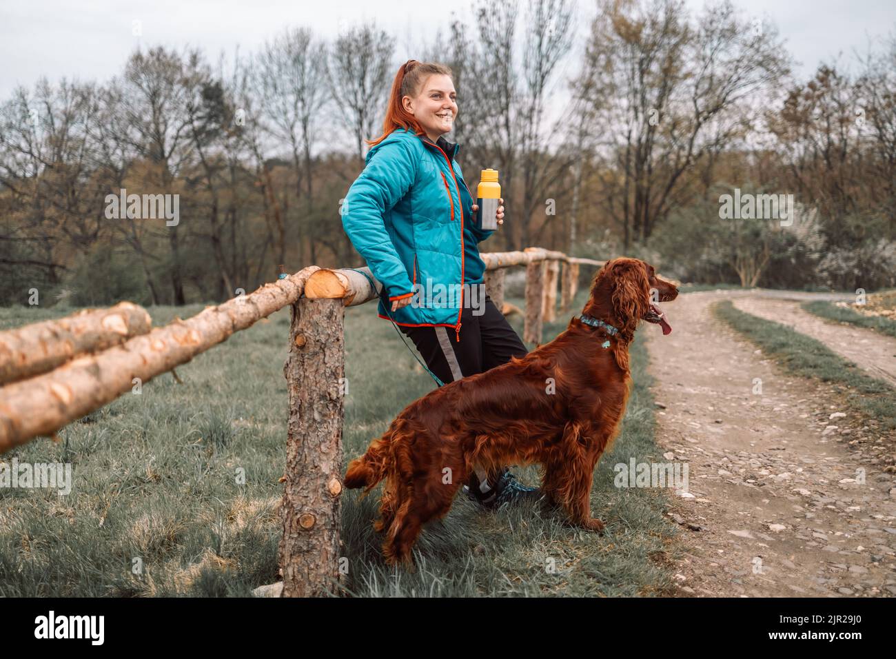 Junge, schöne Sportlerin mit irischem Setter-Hund läuft die Straße entlang und hält Kaffee in einer Tasse Thermobecher. Stockfoto