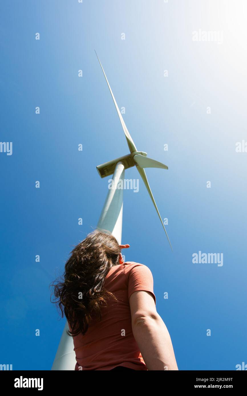 Frau, die an einem sonnigen Tag mit blauem Himmel auf die Windenergieanlage schaut Stockfoto