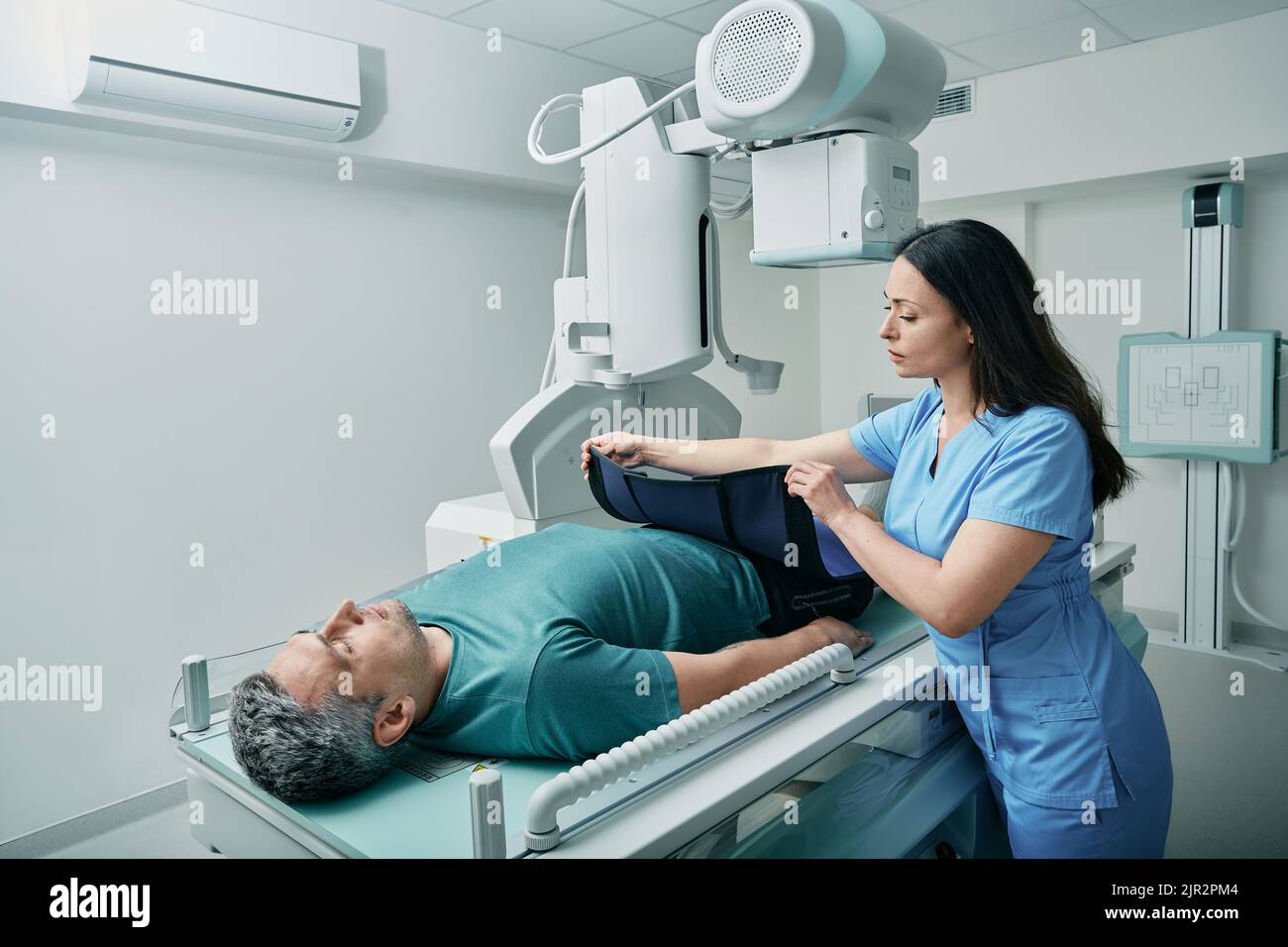 Eine Technikerin bereitet den Patienten im Röntgenraum auf die Körperröntgenuntersuchung vor und stellt ihm eine Strahlenschutzschürze zur Verfügung Stockfoto