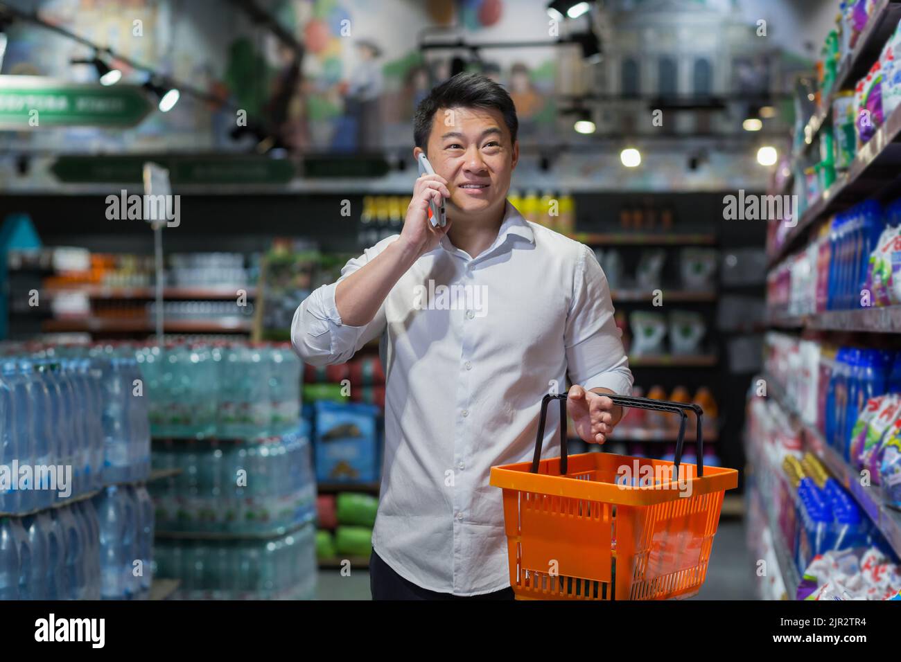 Ein männlicher Käufer in einem Supermarkt wählt Waren aus und kauft ein, ein asiatischer Mann spricht fröhlich am Telefon, konsultiert sich zum Einkaufen Stockfoto