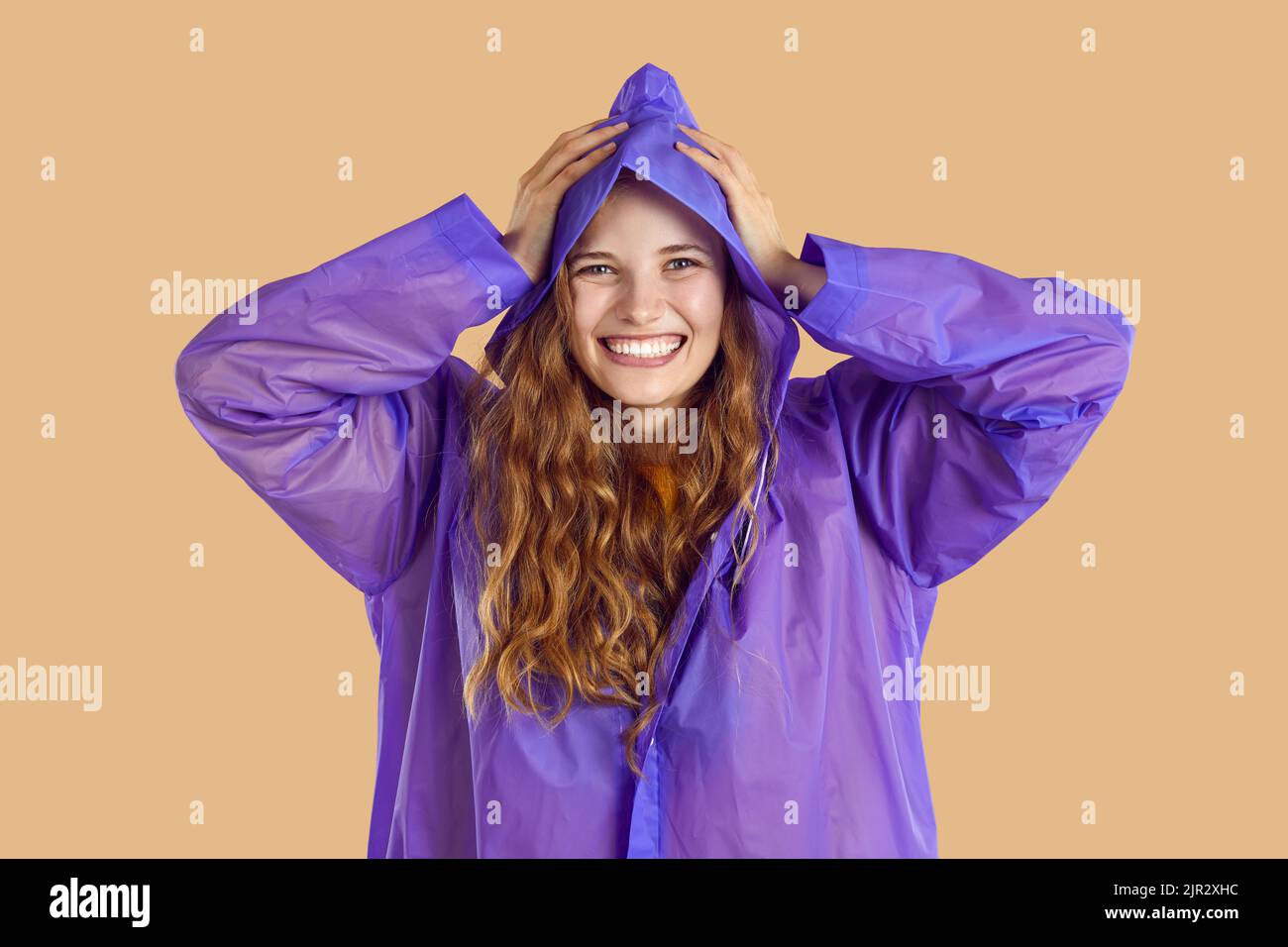 Fröhliche, fröhliche, hübsche junge Frau in einem lila Regenmantel, die sich beim Regen amüsieren kann Stockfoto