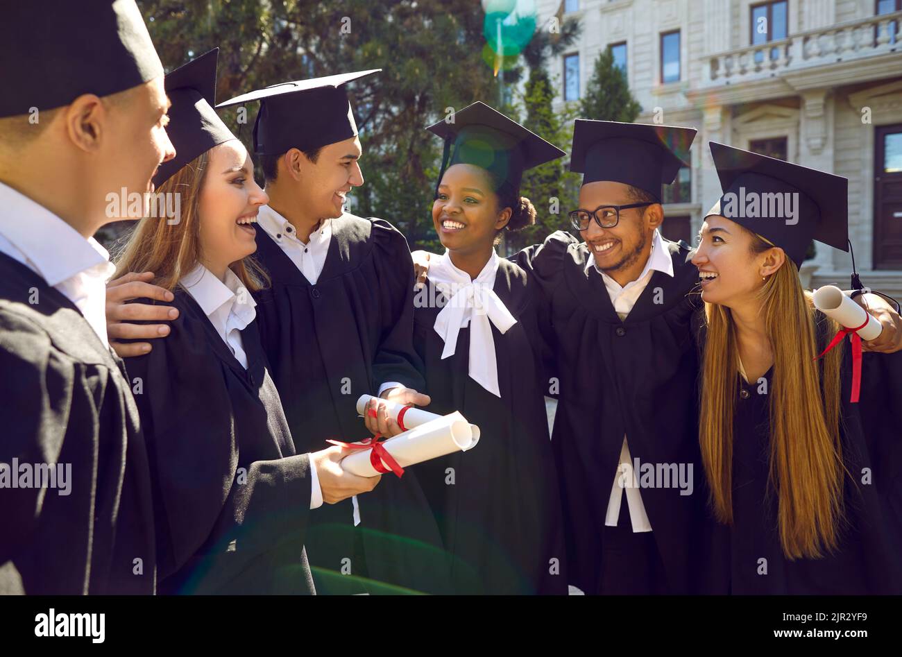 Glückliche multirassische Universitätsabsolventen gehen, reden, lachen und umarmen sich im Freien. Stockfoto