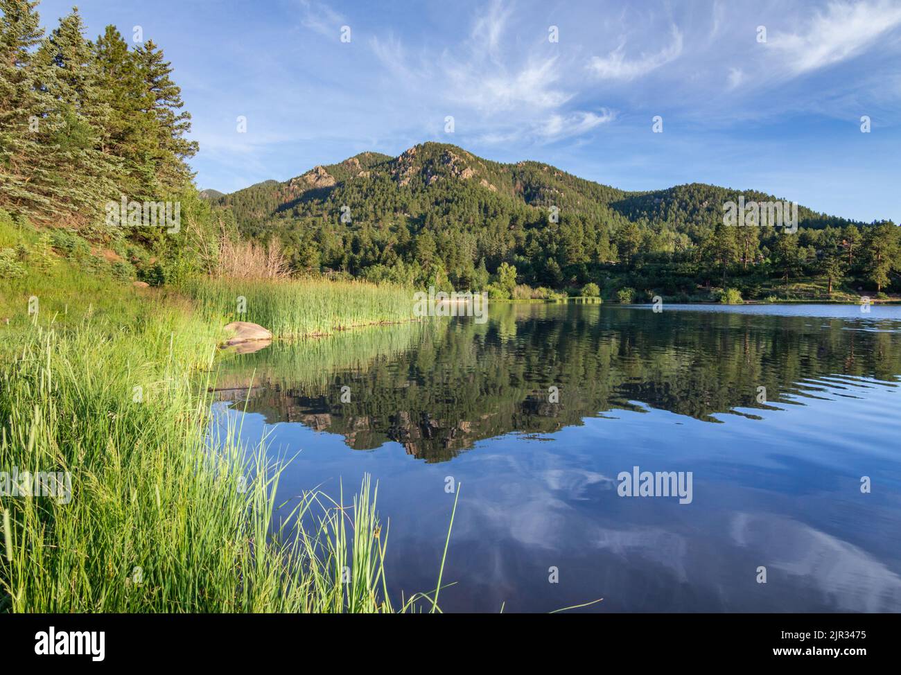 Wispige Wolken und bewaldete Hügel, die sich in ruhigen Gewässern spiegeln, signalisieren einen schönen Start in einen Sommertag in den Rockies Stockfoto