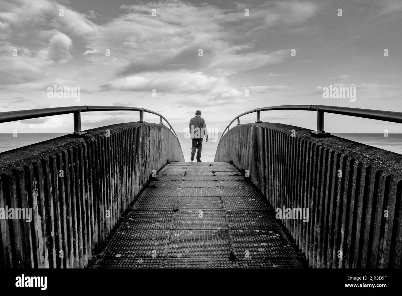 Ein monochromes Bild des Rückens eines Fußgängers, der über die Rainbow Bridge in Colwyn Bay, Nordwales, geht. Das Meer ist im Hintergrund zu sehen. Stockfoto