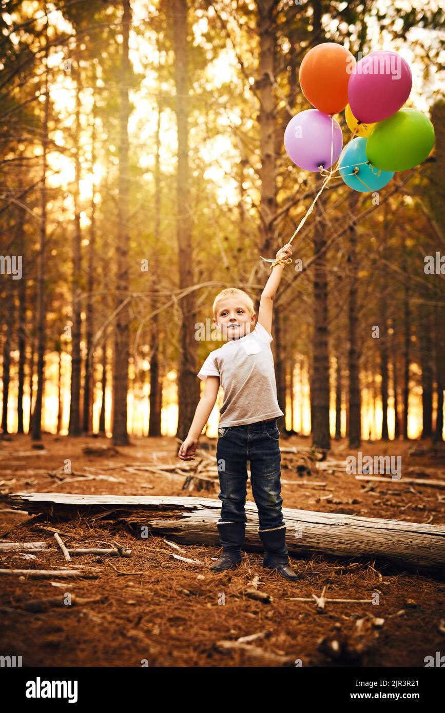 Ein glücklicher kleiner Junge, der einen Haufen Ballons hält, während er draußen im Wald steht. Stockfoto