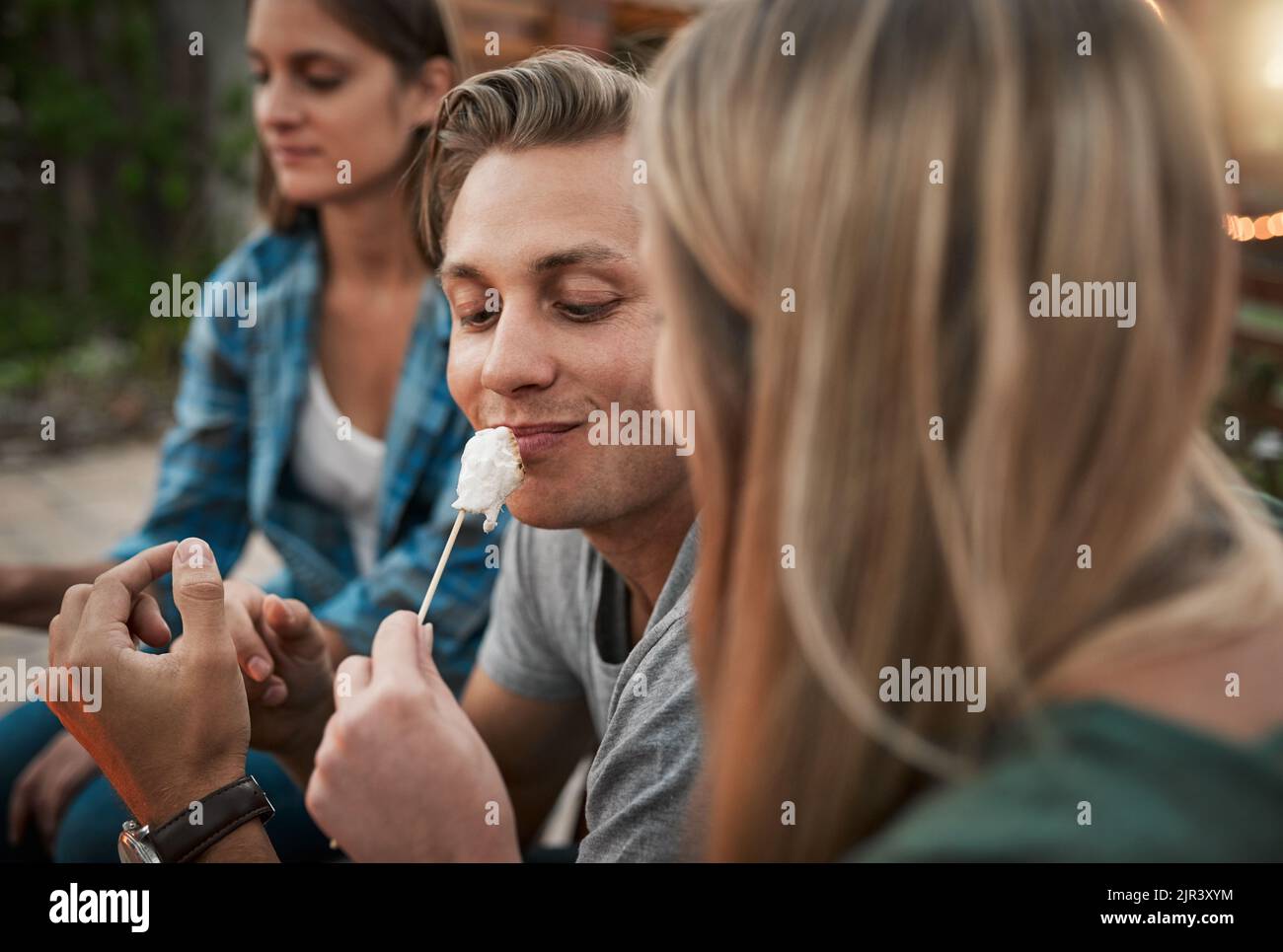 Das ist wirklich lecker. Ein junger, fröhlicher Mann, der von seiner Freundin einen Marschmarschall gefüttert wird, während er draußen sitzt. Stockfoto