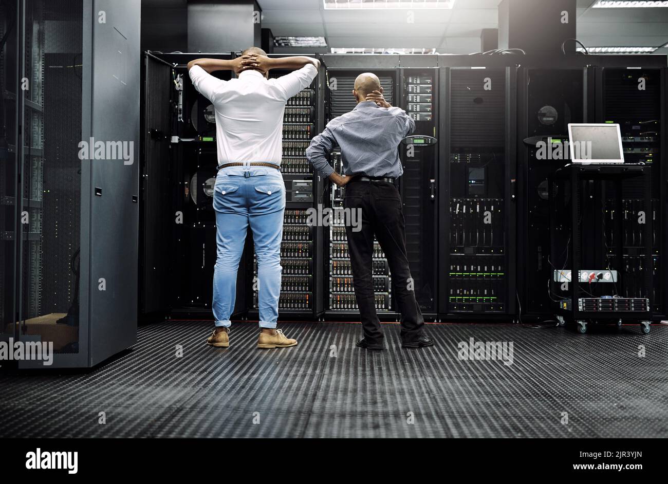 In der Zwischenzeit, im Serverraum... eine Rückansicht von zwei IT-Technikern, die Schwierigkeiten haben, einen Computer in einem Serverraum zu reparieren. Stockfoto