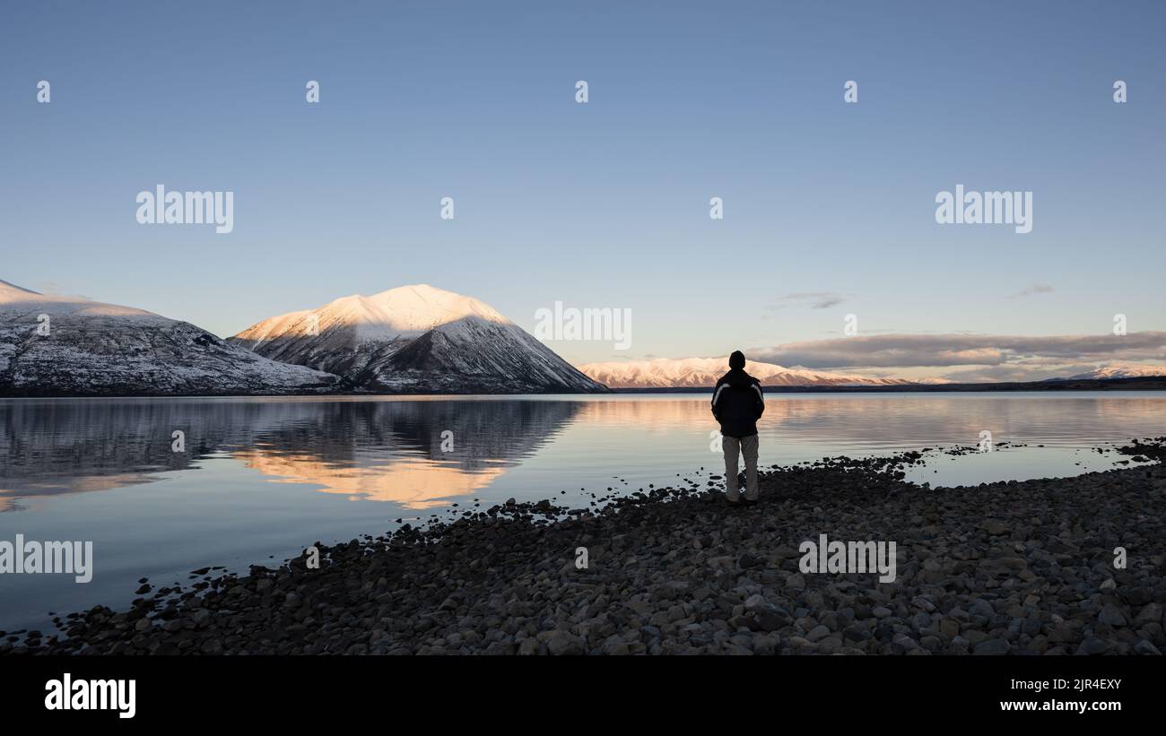Mann, der am Lake Ohau steht und bei Sonnenuntergang auf schneebedeckte Bergketten blickt, Twizel, South Island. Stockfoto