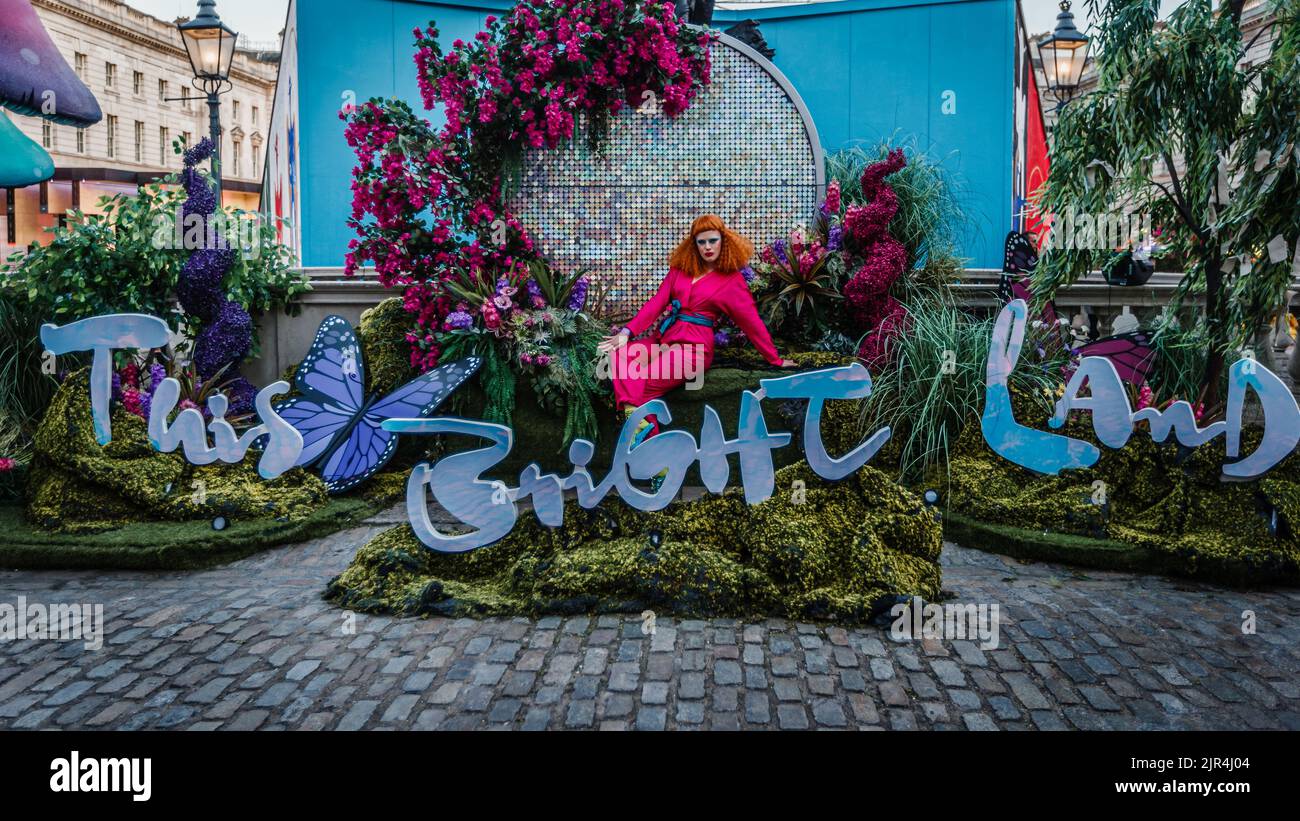 Eine darstellende Künstlerin in einem leuchtend rosa Jumpsuit und einer schockierenden orangen Perücke am Eingang zu The This Bright Land im Somerset House in London. Stockfoto