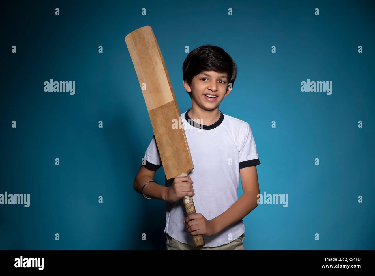 Porträt eines fröhlichen Jungen, der mit einem Cricket-Schläger auf blauem Hintergrund steht Stockfoto