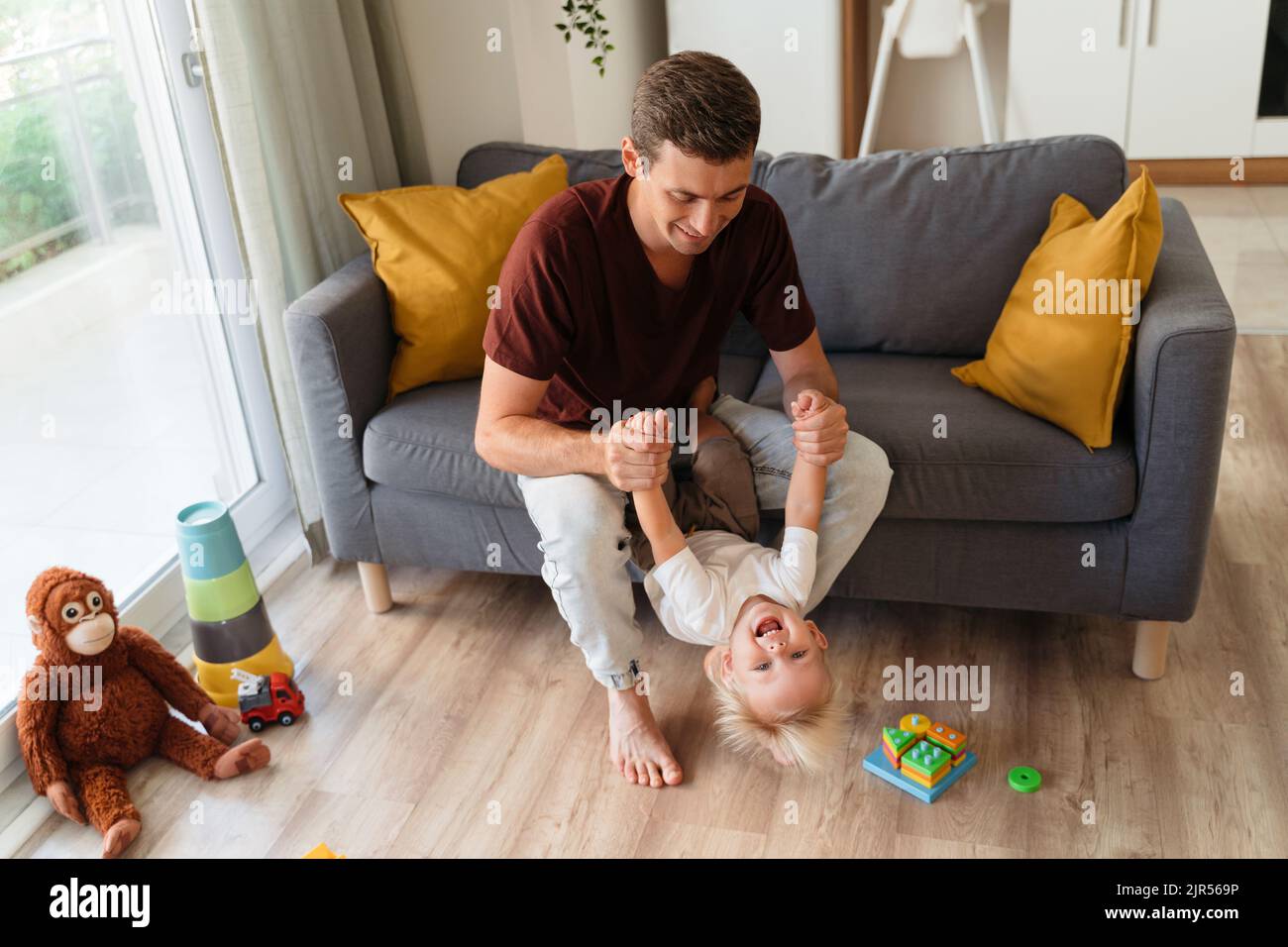 Der junge Vater spielt in aktiven Spielen mit seinem kleinen Jungen, der seine Hände auf dem Sofa im Wohnzimmer zieht, das von Spielzeug auf dem Boden umgeben ist. Glückliche, sorglose Kindheit. Vater-Sohn-Beziehungen, Vaterschaft Stockfoto