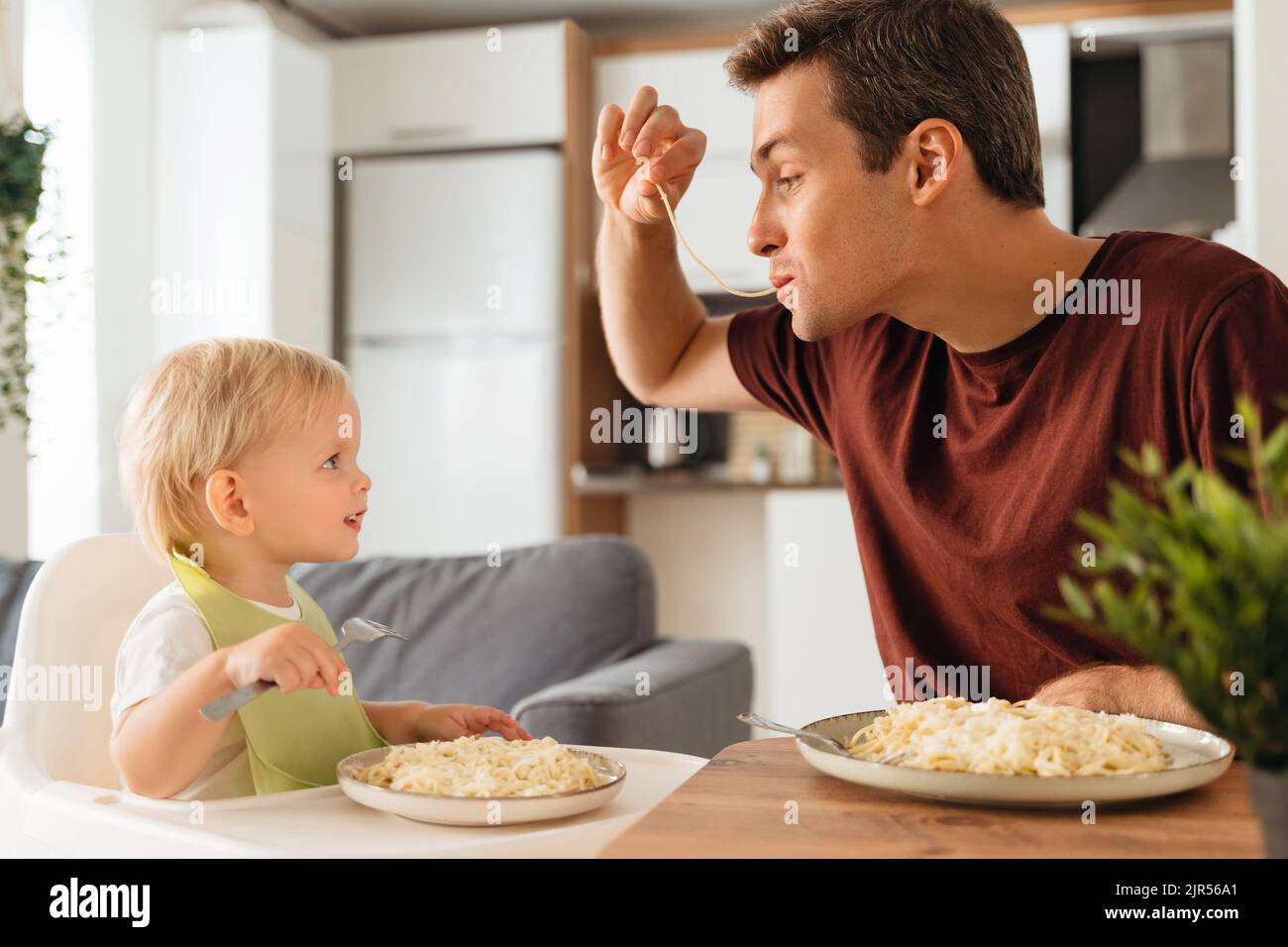 Glücklicher Vater, der mit Spaghetti spielt, während er mit seinem kleinen Jungen im Lätzchen zu Abend isst und dabei mit Gabel isst und lustigen Vater mit Interesse anschaut. Glückliche Vaterschaft, Kind füttern. Vatertag, Familie Stockfoto