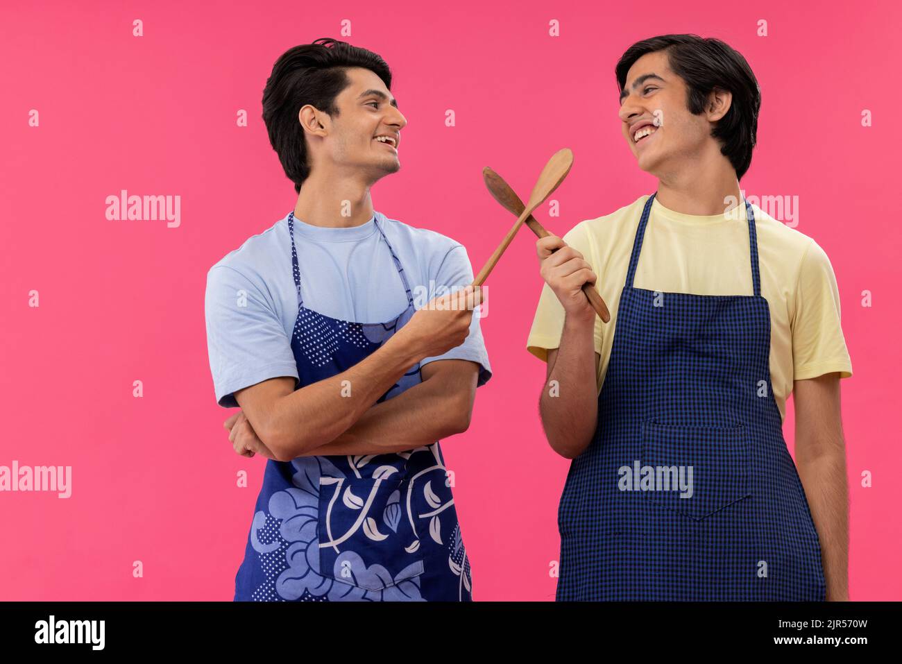 Zwei Jungen im Teenageralter auf der Kochschürze, die mit Spateln vor rosafarbenem Hintergrund stehen Stockfoto