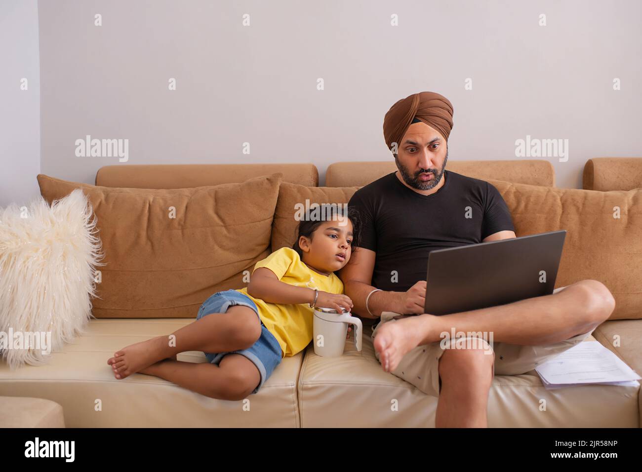 Sikh-Mann, der am Laptop arbeitete, während die Tochter neben ihm auf dem Sofa lag Stockfoto