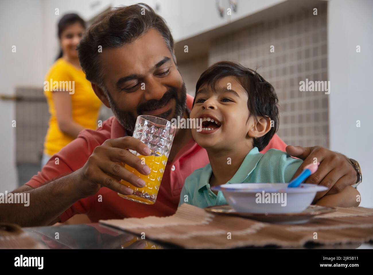 Nahaufnahme des fürsorglichen Vaters, der seinem kleinen Kind und seiner Mutter, die dahinter steht, Orangensaft zufüttert Stockfoto