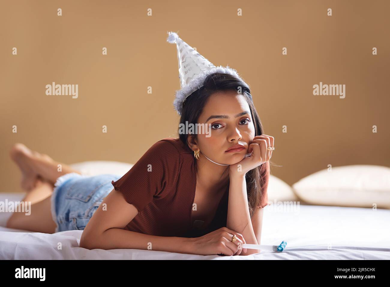 Traurige junge Frau mit Geburtstagsmütze, die auf dem Bett liegt und ihr Kinn auf der Hand liegt Stockfoto