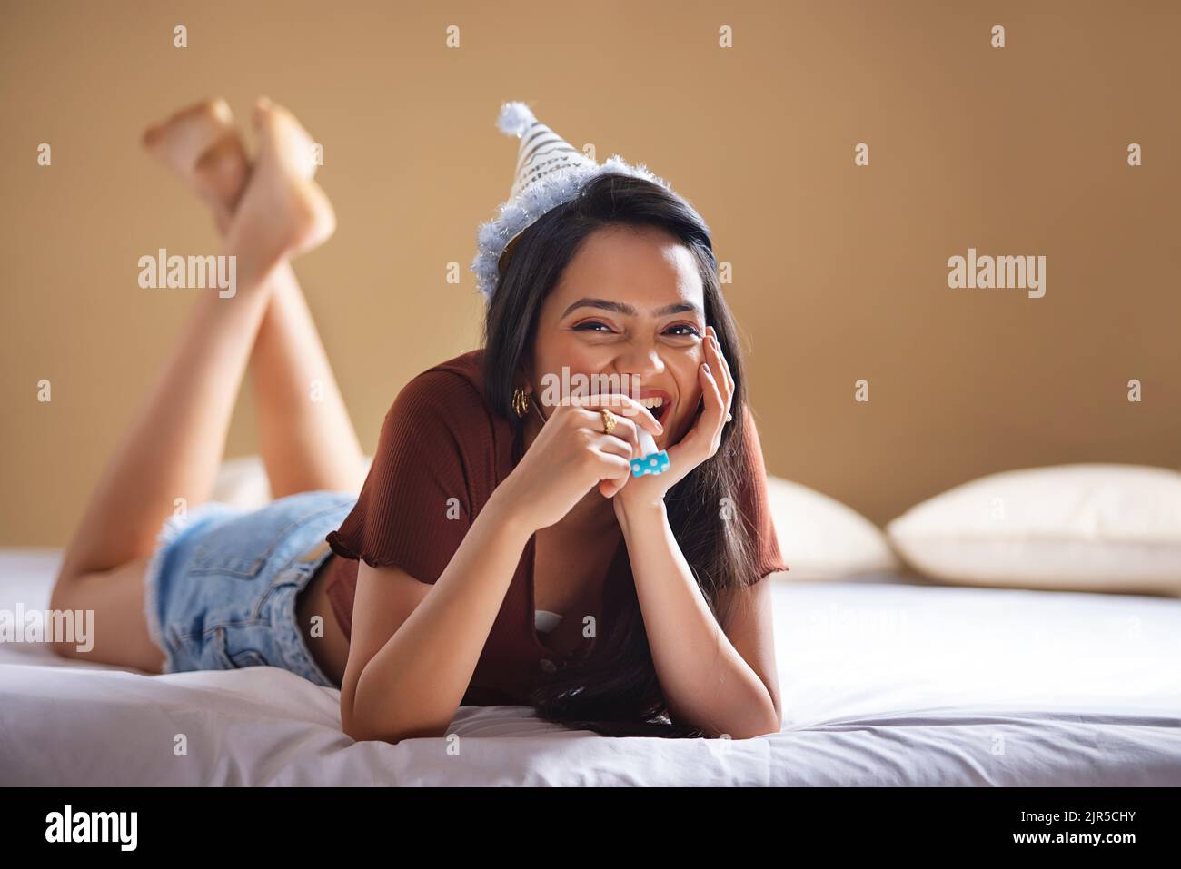Glückliche junge Frau mit Geburtstagshut, die auf dem Bett liegt und ihr Kinn auf der Hand liegt Stockfoto