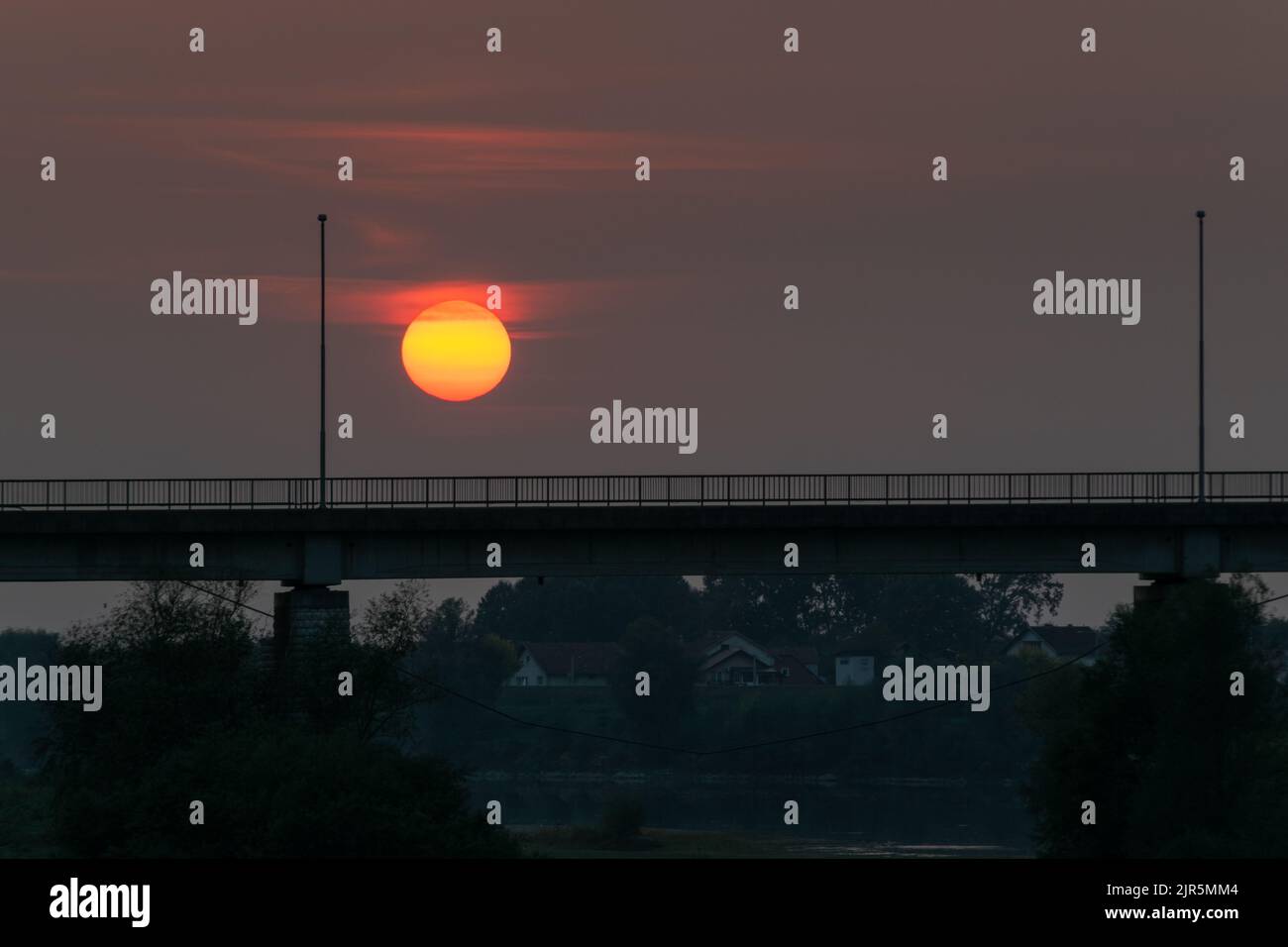 Silhouette der Brücke auf dem Fluss Sava zwischen Bosnien und Herzegowina und Kroatien gegen einen bewölkten Himmel mit einer großen orangefarbenen Sonne Stockfoto