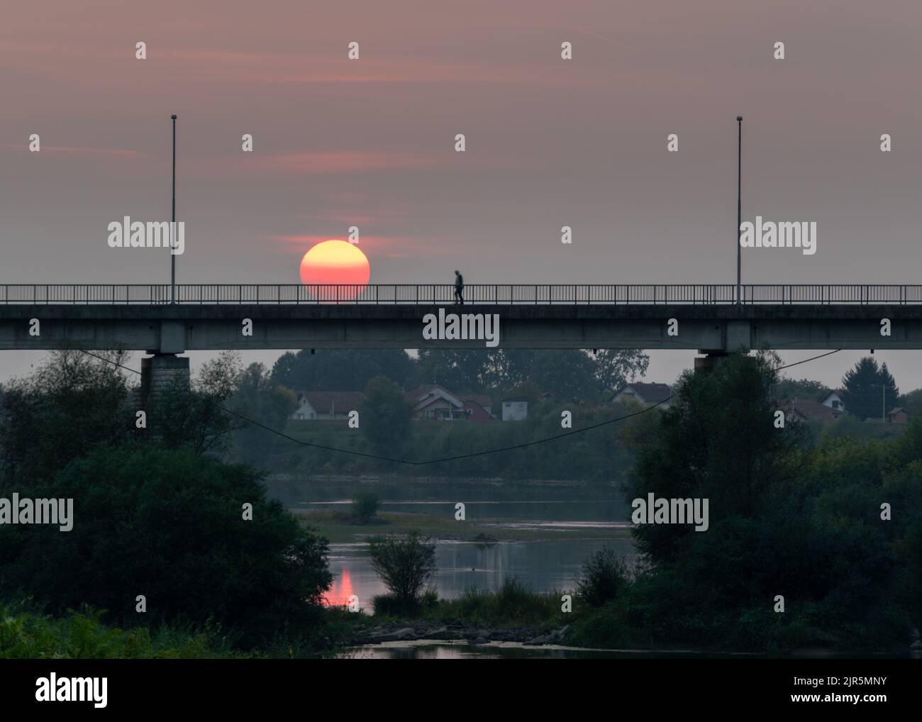 Ein Mann geht auf einer Brücke über den Fluss Sava zwischen Bosnien und Herzegowina und Kroatien gegen einen bewölkten Himmel mit einer großen roten Sonne. Stockfoto
