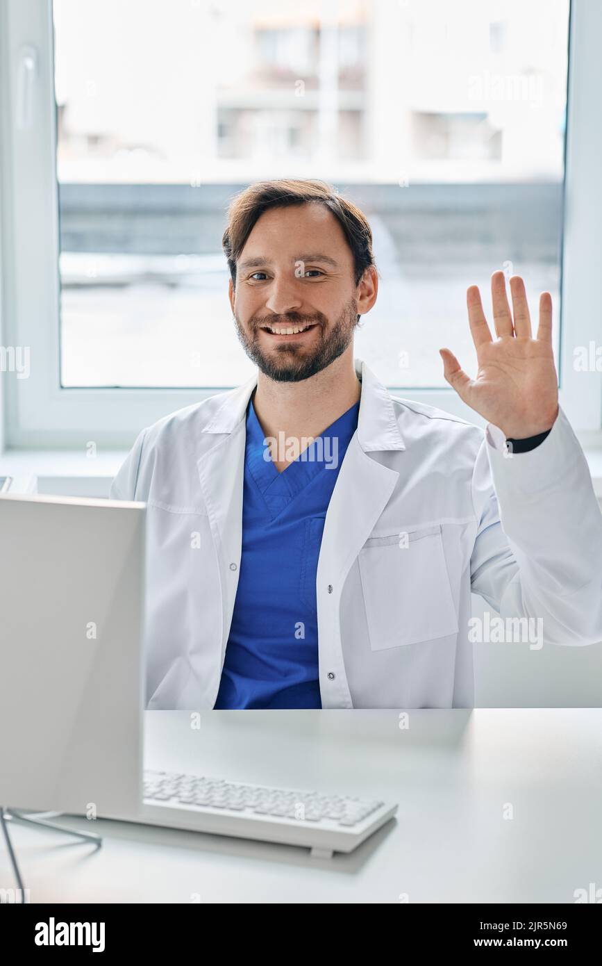 Freundlicher Arzt, der an seinem Arbeitsplatz in einer medizinischen Uniform sitzt und im Krankenhaus mit der Hand winkt Stockfoto