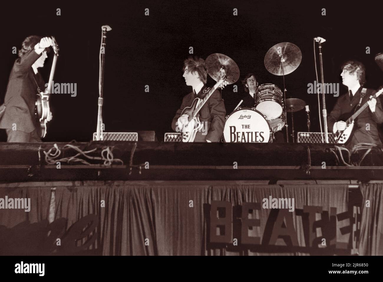 Die Beatles auf der Bühne beim Gator Bowl in Jacksonville, Florida, am 11. September 1964. Hurrikan Dora hatte Jacksonville am Vortag getroffen, und die verbleibenden starken Winde, die auf 45 mph zustürmen, erforderten, dass Ringo Starr's Trommeln auf die Bühne genagelt wurden und dass der Pappschriftzug der Beatles auf der Seite der Bühne schließlich weggerissen wurde. (USA) Stockfoto