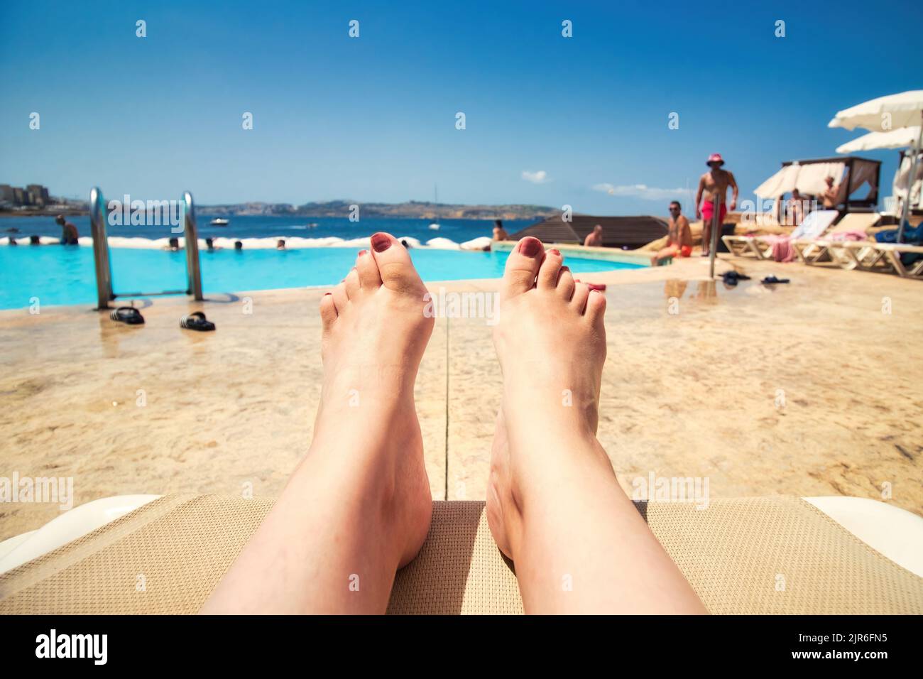 Aus der Sicht der ersten Person von einer Frau, die entspannt auf einer Sonnenliege am Pool liegt und auf den Pool blickt, mit Menschen im Hintergrund und Copy-SP Stockfoto