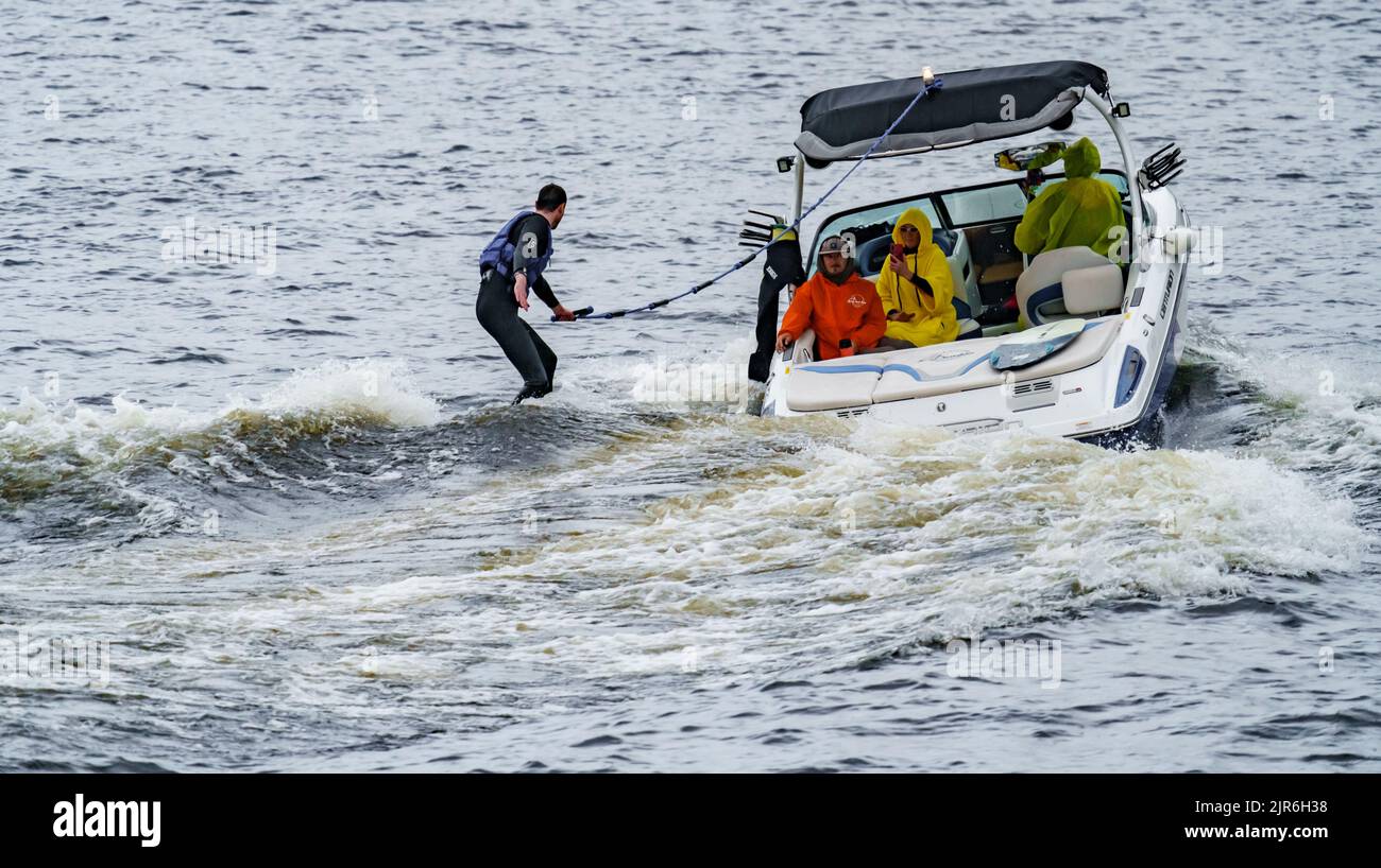 Russland, St. Petersburg, 29. Juli 2022: Mann auf Wakeboard auf Welle des Motorbootes im Sommerfluss, Wakesurfen auf Fluss Sommerferien Stockfoto