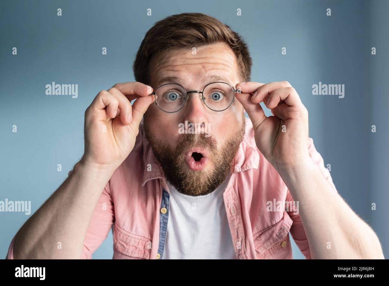 Der Mensch ist schockiert, er glaubt seinen Augen nicht, er hält eine Brille mit den Händen und öffnet überraschend den Mund. Blauer Hintergrund. Stockfoto