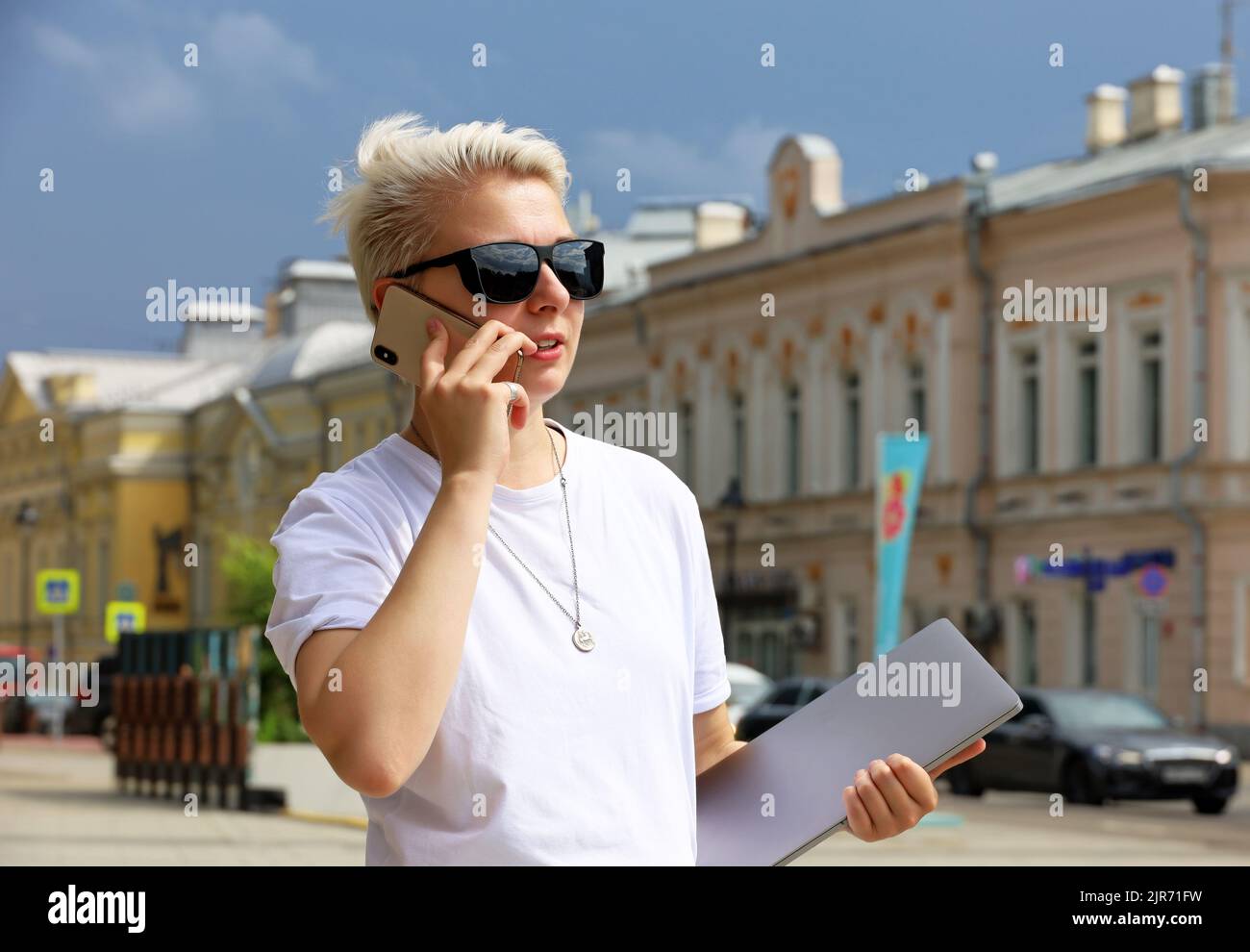 Mädchen mit kurzen blonden Haaren in Sonnenbrillen, die auf dem Mobiltelefon sprechen, während sie ihren Laptop in den Händen auf der Stadtstraße halten. Tomboy Lifestyle im Sommer Stockfoto