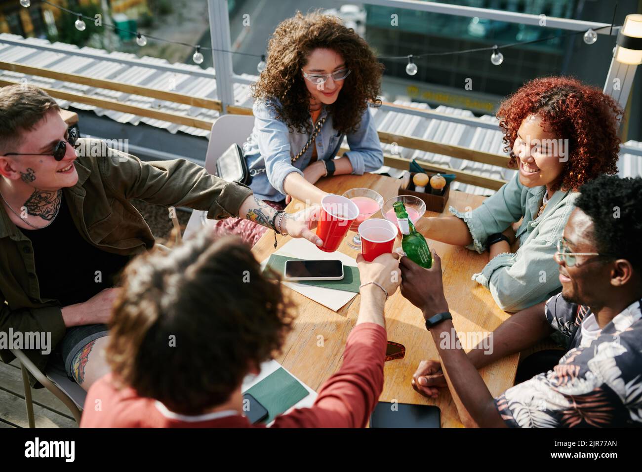 Fünf fröhliche Jugendliche Freunde klirren mit ihren Getränken über dem Tisch, während sie sich am sonnigen Sommertag im Café ausruhen Stockfoto