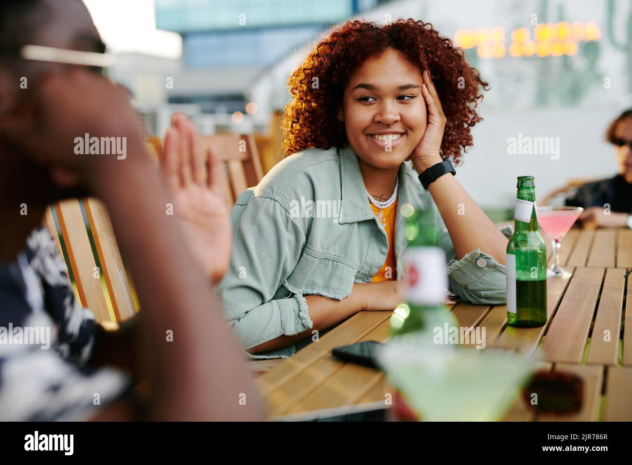 Glückliche junge schwarze Frau in Casualwear, die ihren Freund lächelnd ansieht, während sie vor ihm am Tisch sitzt und Bier trinkt Stockfoto