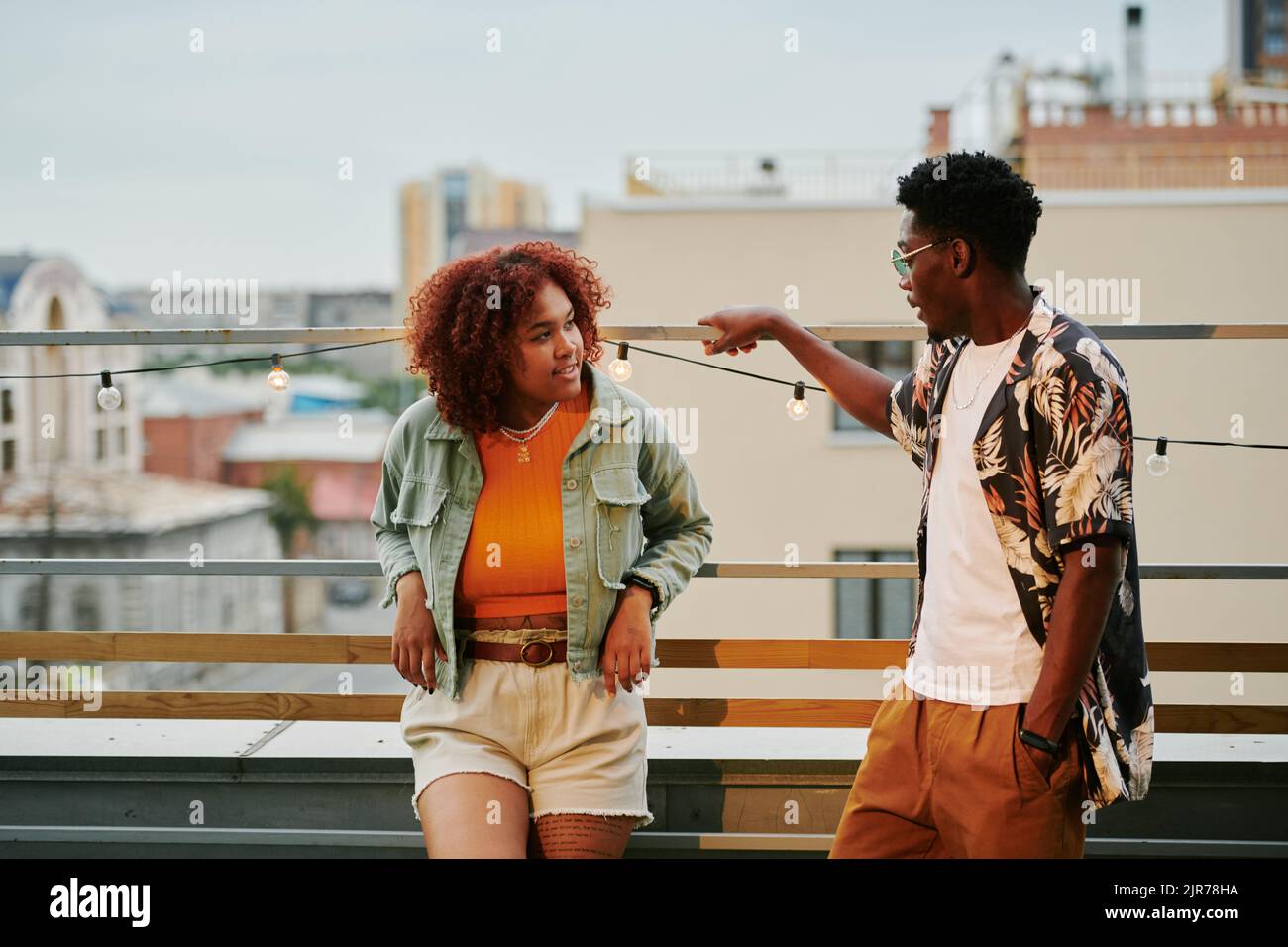 Junges afroamerikanisches Paar in stilvoller Casualwear, das sich auf der Terrasse des Dachcafés gegen die städtische Umgebung unterhielt Stockfoto