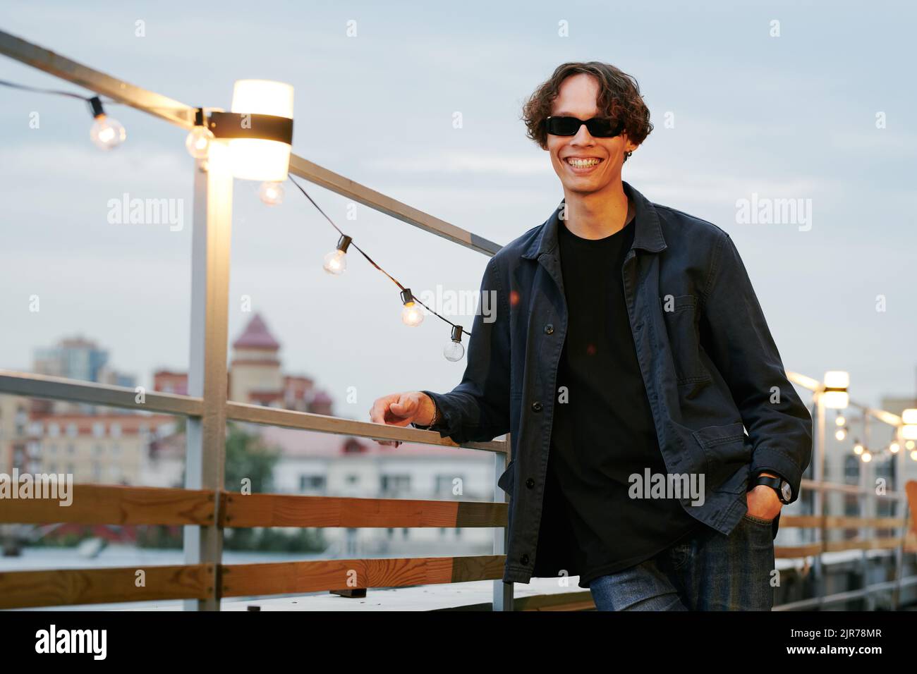 Fröhlicher Mann mit einem toothy Lächeln, der an der Beleuchtung hing, während er die Dachparty im Café im Freien genoss Stockfoto