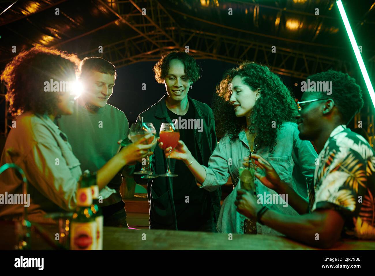 Fünf fröhliche Jugendliche interkulturelle Menschen in stilvoller Casualwear, die bei Cocktails anklirren, während sie auf der Party vor einander stehen Stockfoto