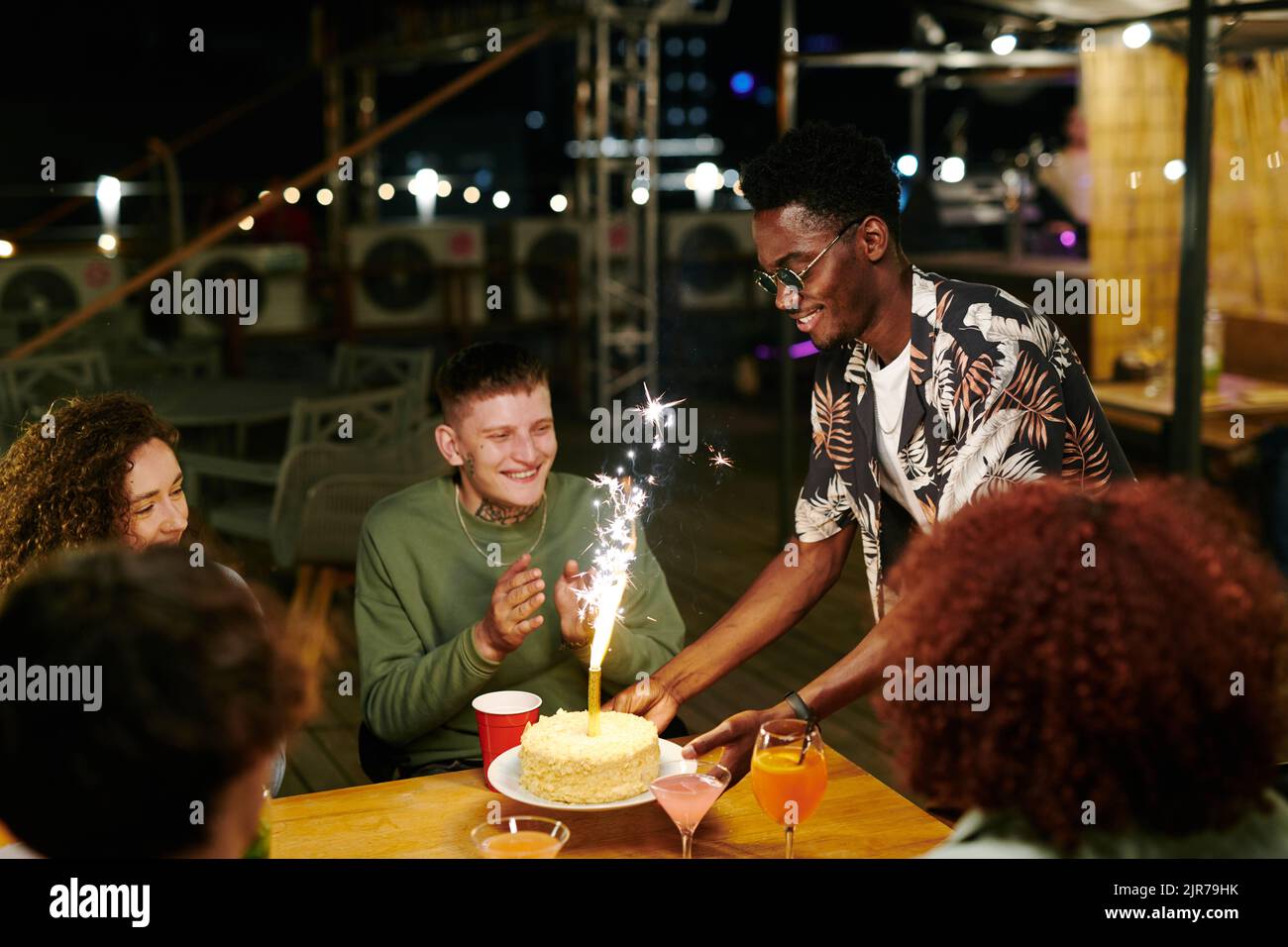 Fröhlicher junger afroamerikanischer Mann, der Teller mit leckerem Kuchen und Glitzer auf einen festlichen Tisch legte, an dem sich seine Freunde zur Party versammelten Stockfoto
