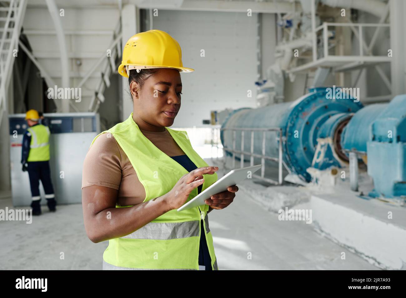 Junge ernsthafte schwarze Frau mit Tablet, die technische Online-Daten durchschaut, während sie vor der Kamera in der Werkstatt der Fabrik steht Stockfoto