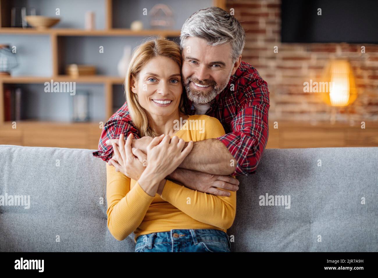 Fröhlicher, erwachsener kaukasischer Mann umarmte Frau, genoss einen zarten Moment und schaute in der Freizeit auf die Kamera Stockfoto