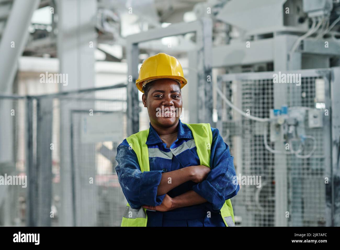 Junge erfolgreiche afroamerikanische Arbeiterin der Fabrik in Uniform und Schutzhelm, die Sie während des Stehens in der Werkstatt anschaut Stockfoto