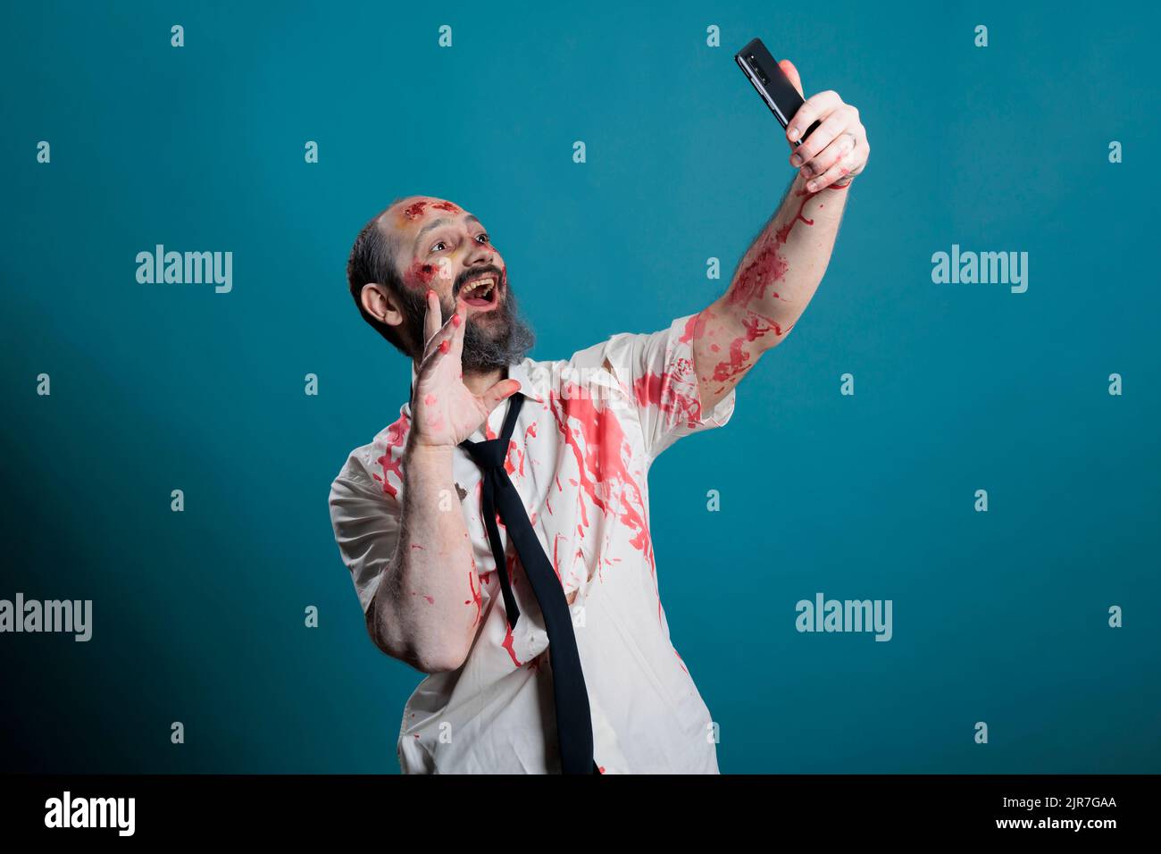 Apokalyptische Leiche, die auf dem Smartphone fotografiert, sich vor der Kamera herumtollt und unheimlich agiert. Gruseliger Zombie mit hässlichen Wunden und blutigen Narben, die Fotos auf dem Handy machen, Hirnessen. Stockfoto