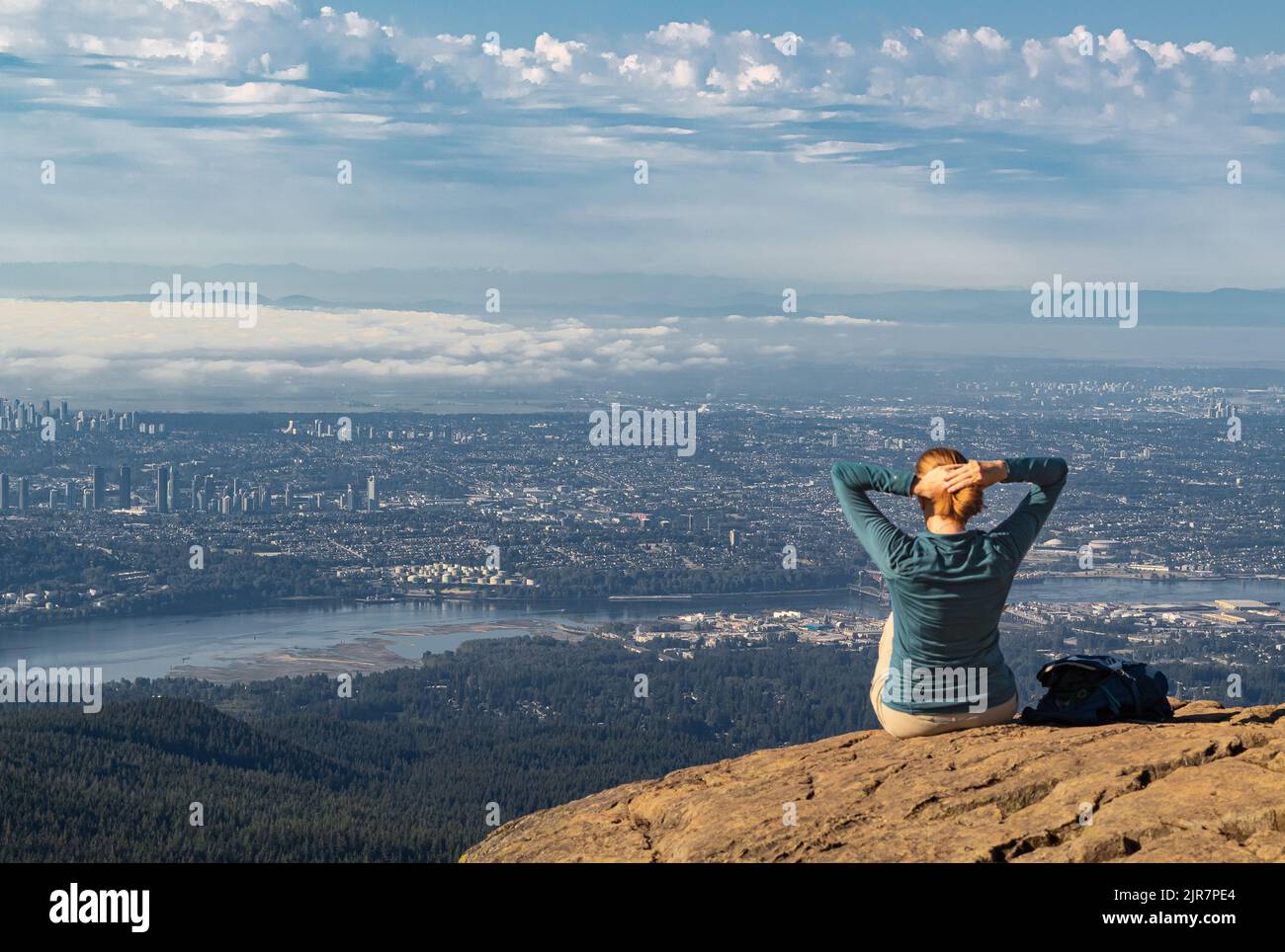 Aktive Frau, die am Rand der Klippe sitzt und den Blick auf das Tal, den Fluss und die Berge genießt. Reisen entlang Kanadas Stockfoto