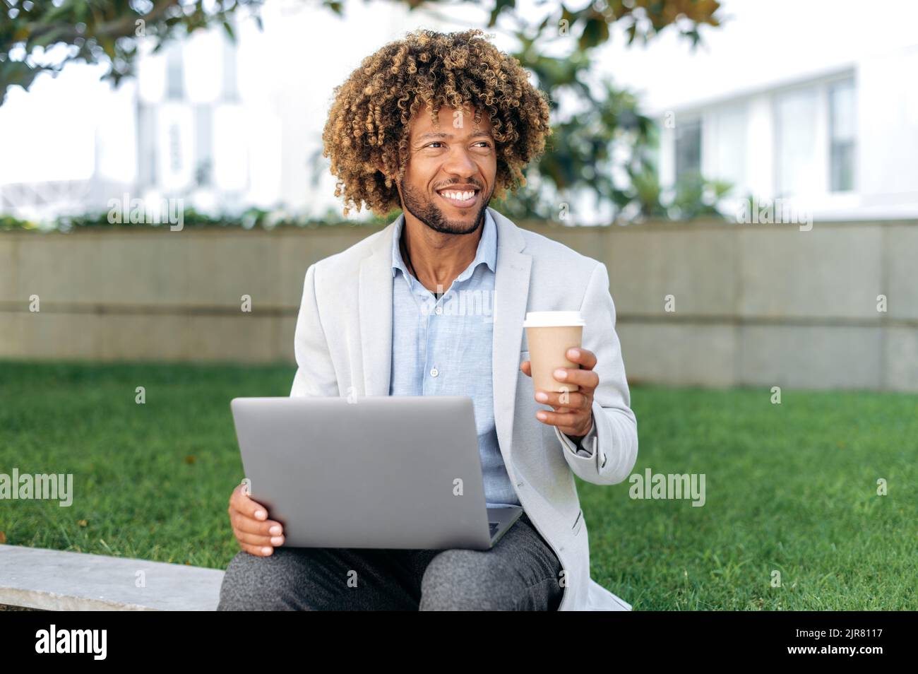 Positiv schöner erfolgreicher brasilianischer oder spanischer Mann, Firmenangestellter, Unternehmer, sitzt im Freien mit Laptop in den Händen, hält ein Glas Kaffee in der Hand, pausiert bei der Arbeit, schaut weg, lächelt Stockfoto