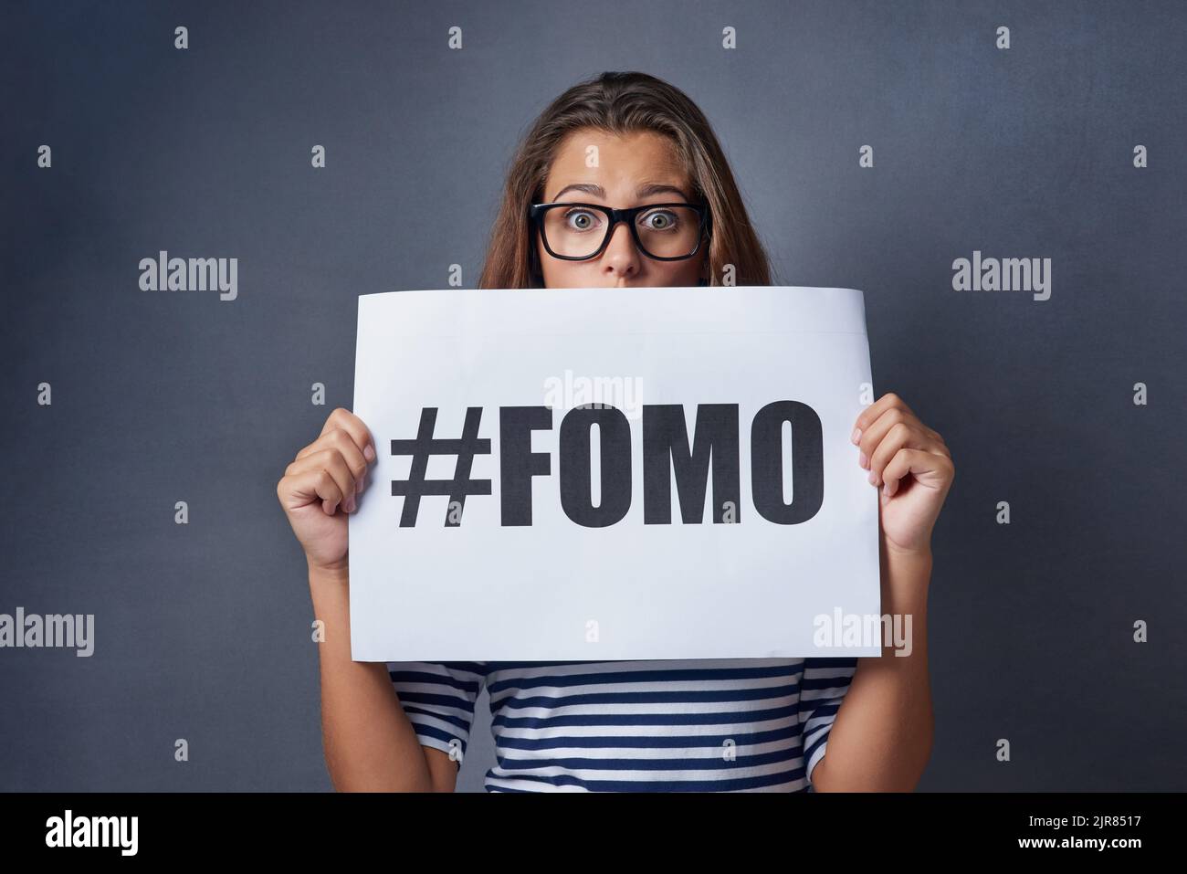 Wenn es online passiert ist, muss es real sein. Studioaufnahme einer attraktiven jungen Frau, die ein Schild mit FOMO-Aufdruck auf grauem Hintergrund hält. Stockfoto